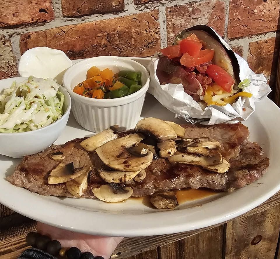 Plate of grilled steak topped with mushrooms, served with coleslaw, mixed vegetables, and a baked potato with salsa