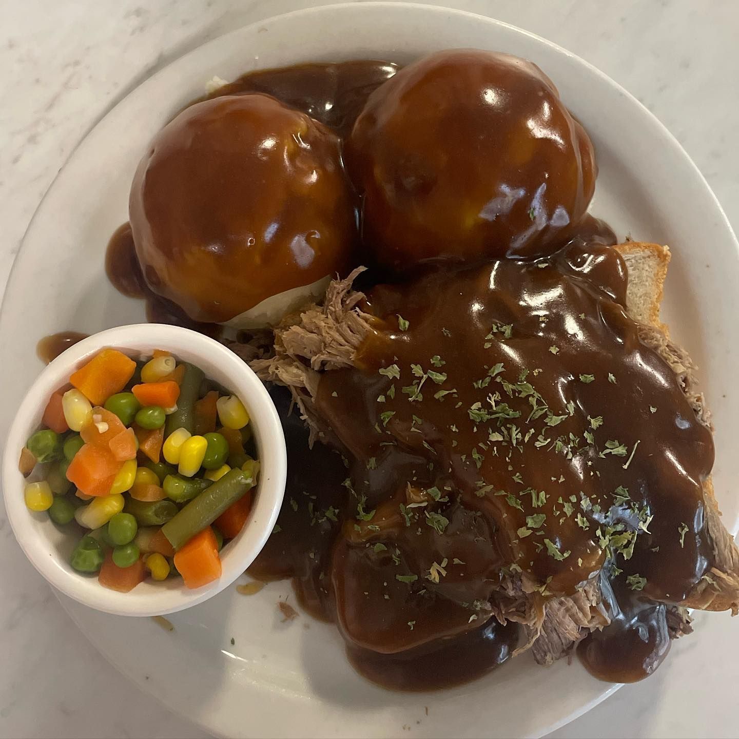 Plated roast dinner with brown gravy, three mashed potatoes, shredded meat, and a cup of mixed vegetables