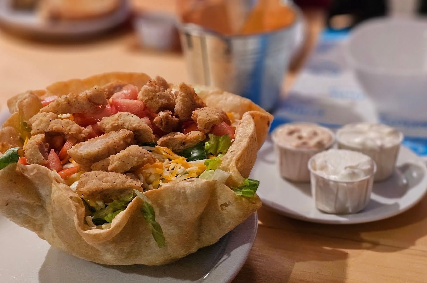 Loaded taco salad in a fried tortilla bowl with dipping sauces on a table