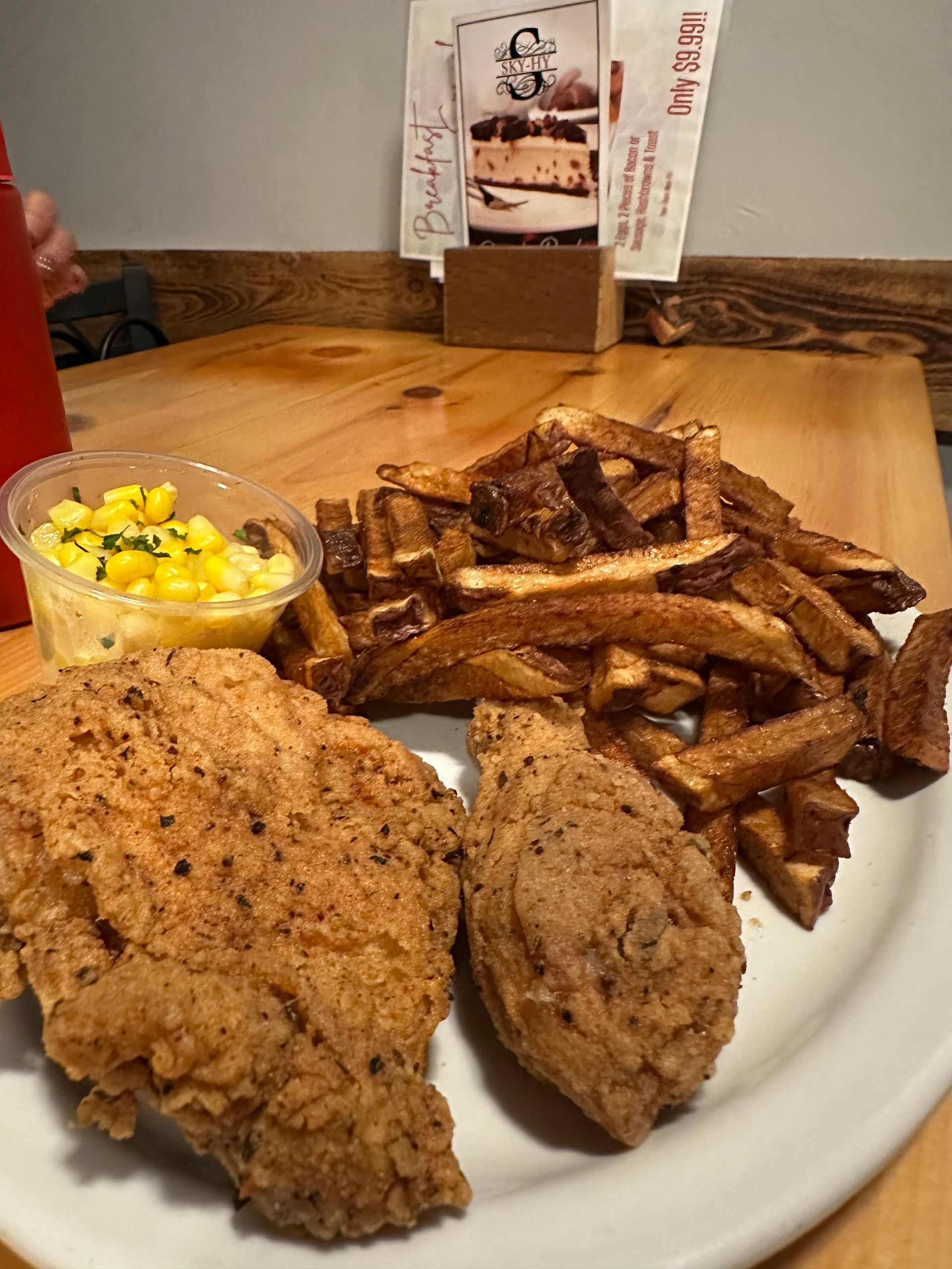 Plate of fried chicken, fries, and corn on a wooden table in a restaurant.