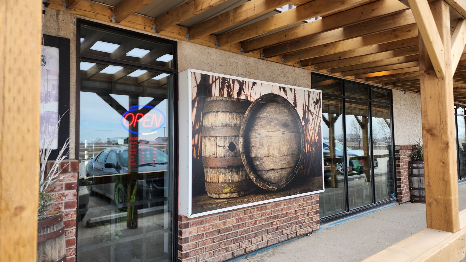 Covered outdoor storefront with wooden beams and a large barrel mural beside glass doors.
