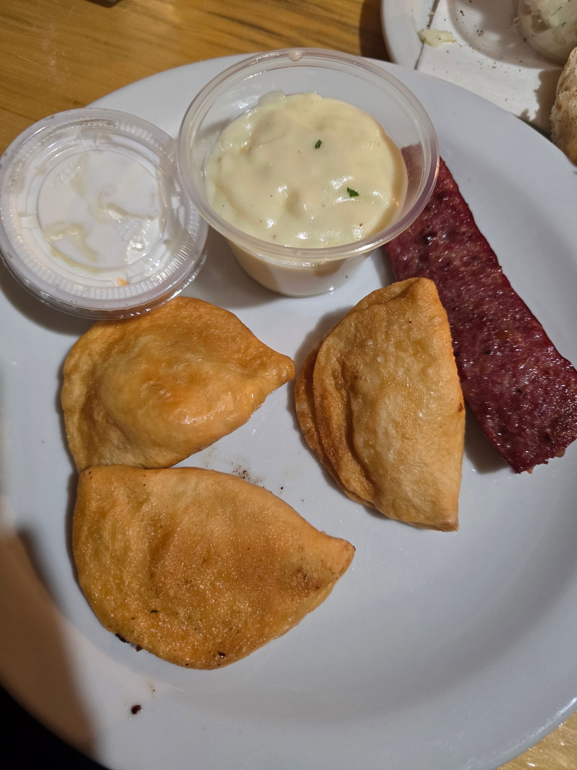 Plate with three fried pastries, a cup of creamy dip, and a strip of cured meat on a white dish.