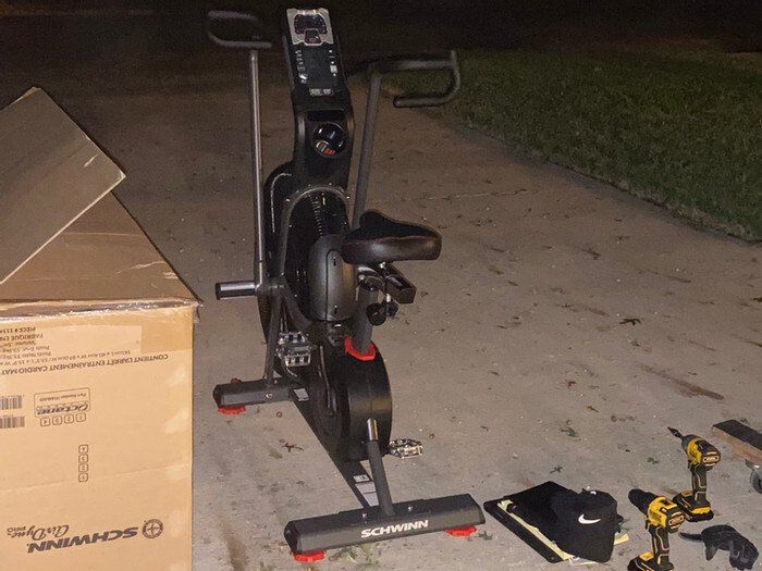 Black stationary exercise bike on a driveway beside a cardboard box, with tools and shoes nearby.
