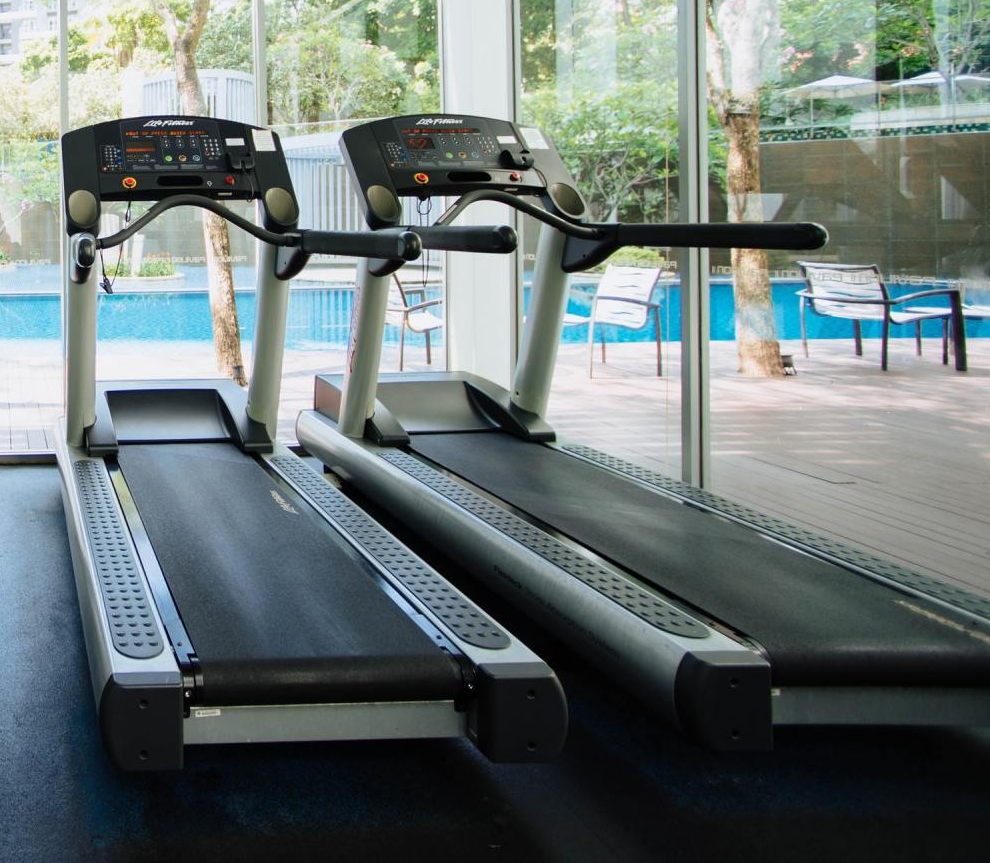 Two treadmills in a bright gym by a pool and glass windows