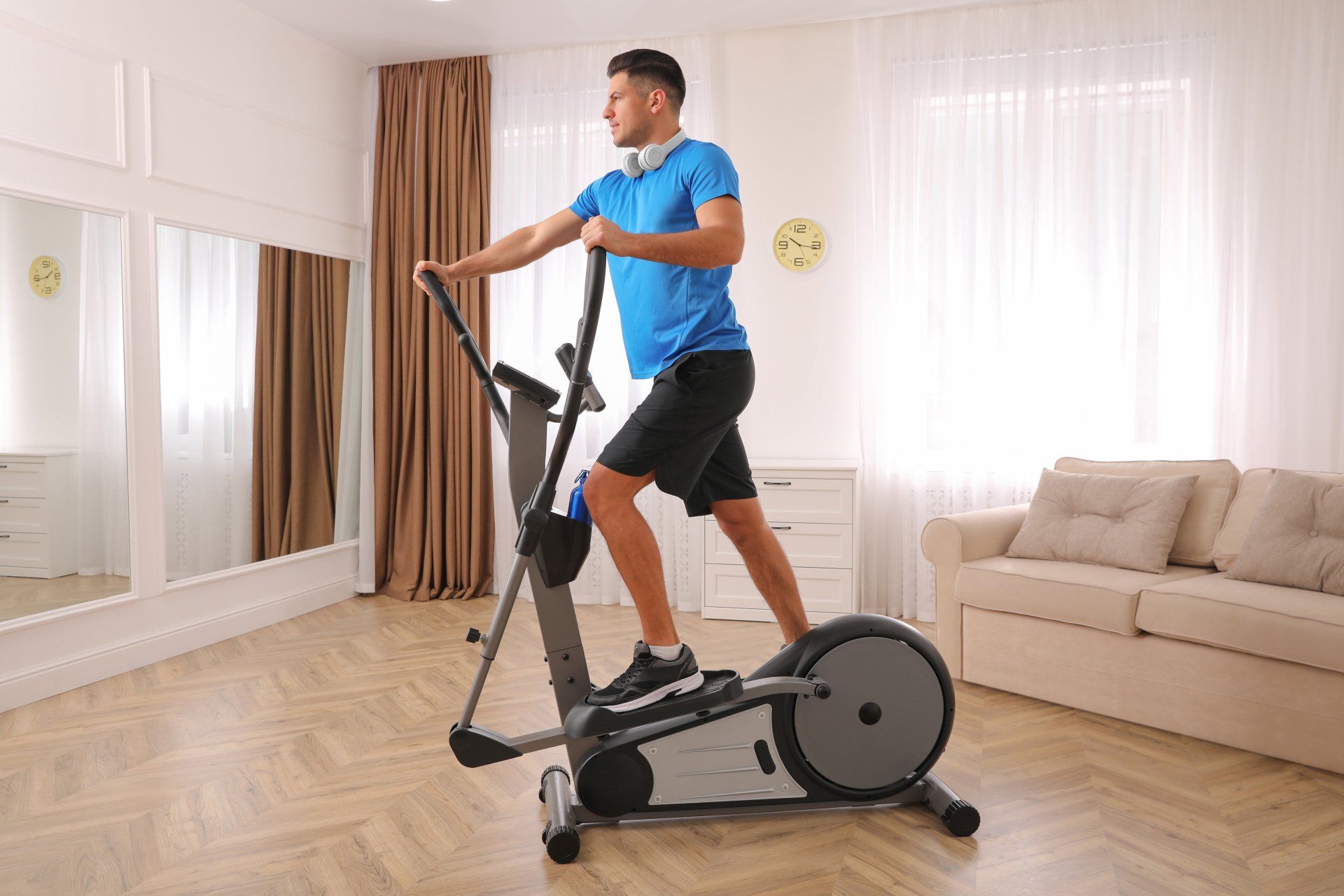 Man using an elliptical machine in a bright living room with beige sofa and mirrors