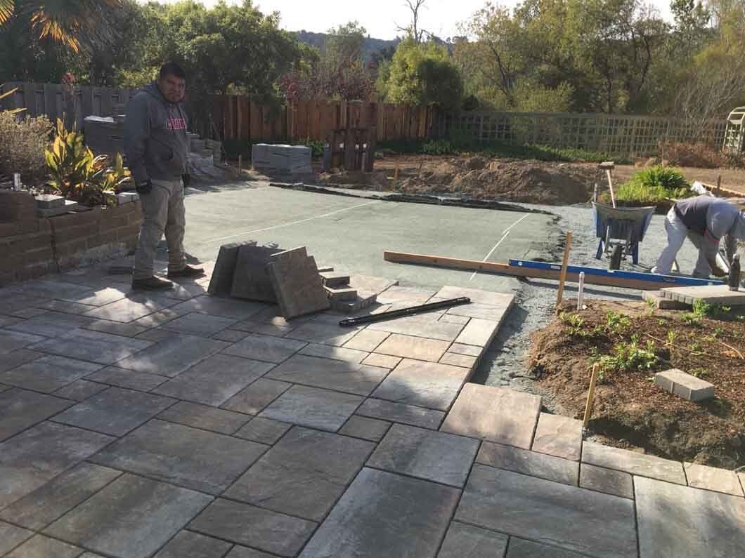 Two men are working on a patio in a backyard.