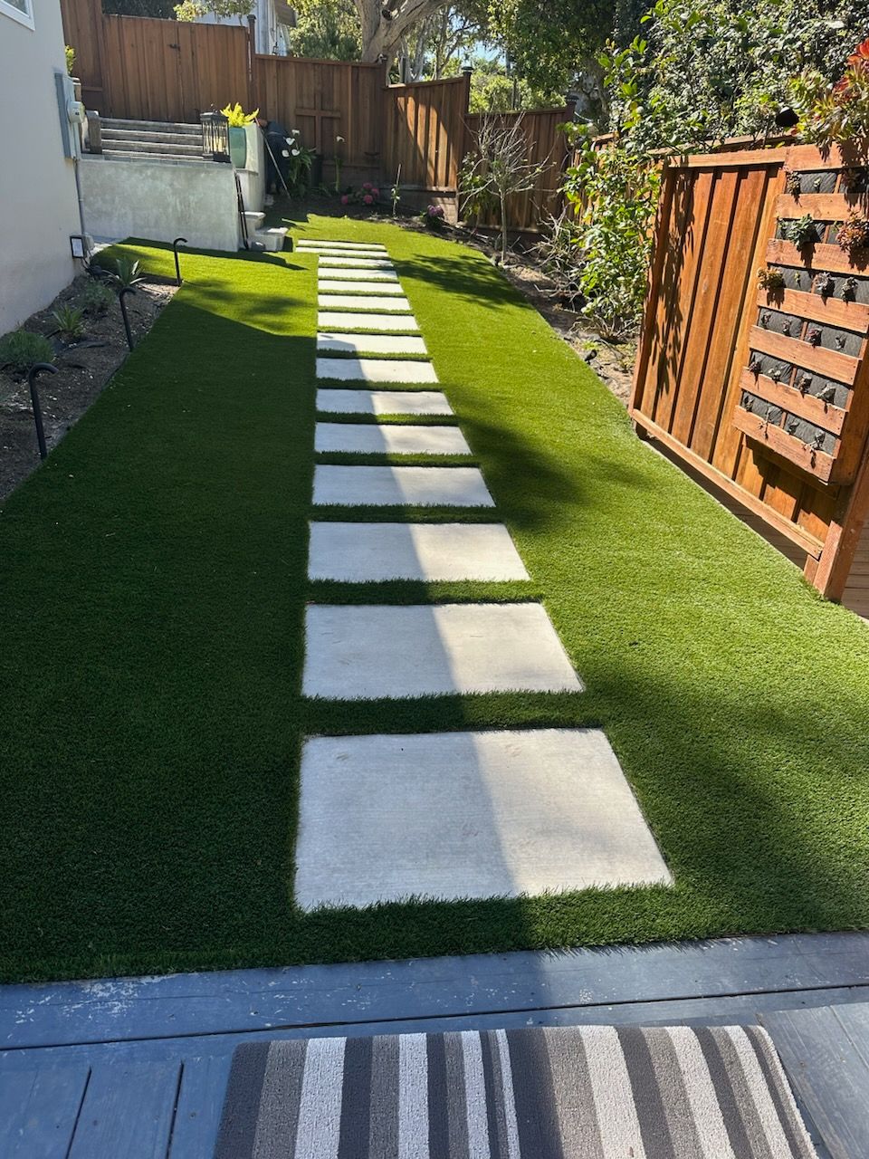 Pathway of square stepping stones through artificial turf. Wooden fence and house in the background.