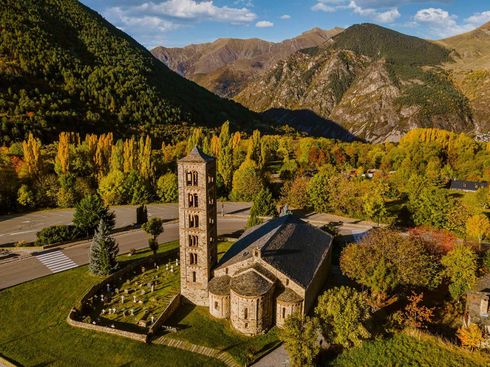 Església de pedra amb un campanar alt en una vall de tardor, envoltada d’arbres daurats i muntanyes