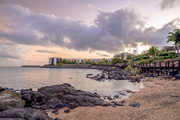 A Beach With Rocks and a Building in the Background at Sunset — Coastal Arborist Services in Whitsundays, QLD