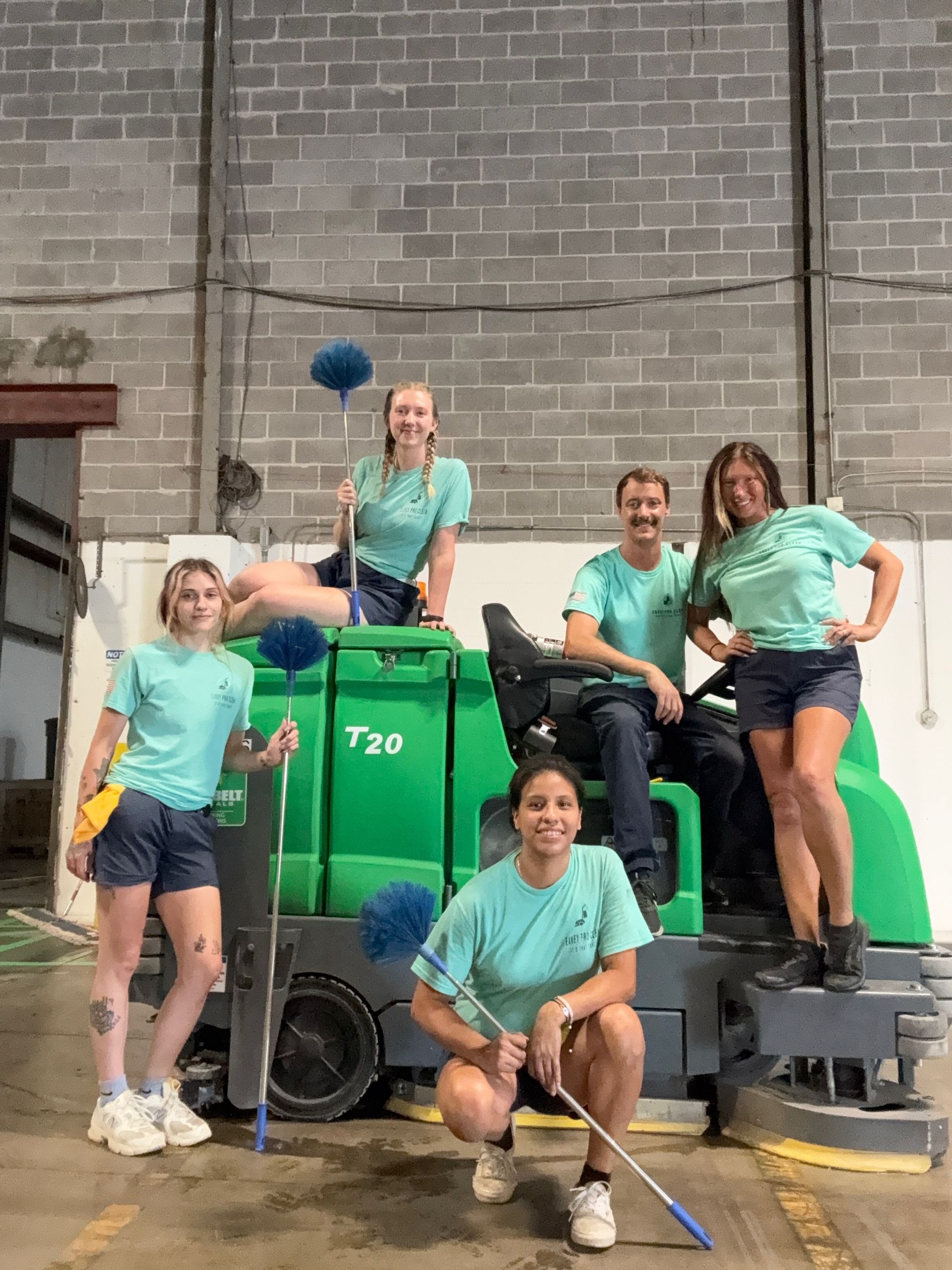 Group of people in matching teal shirts and shorts pose with cleaning equipment in a warehouse.