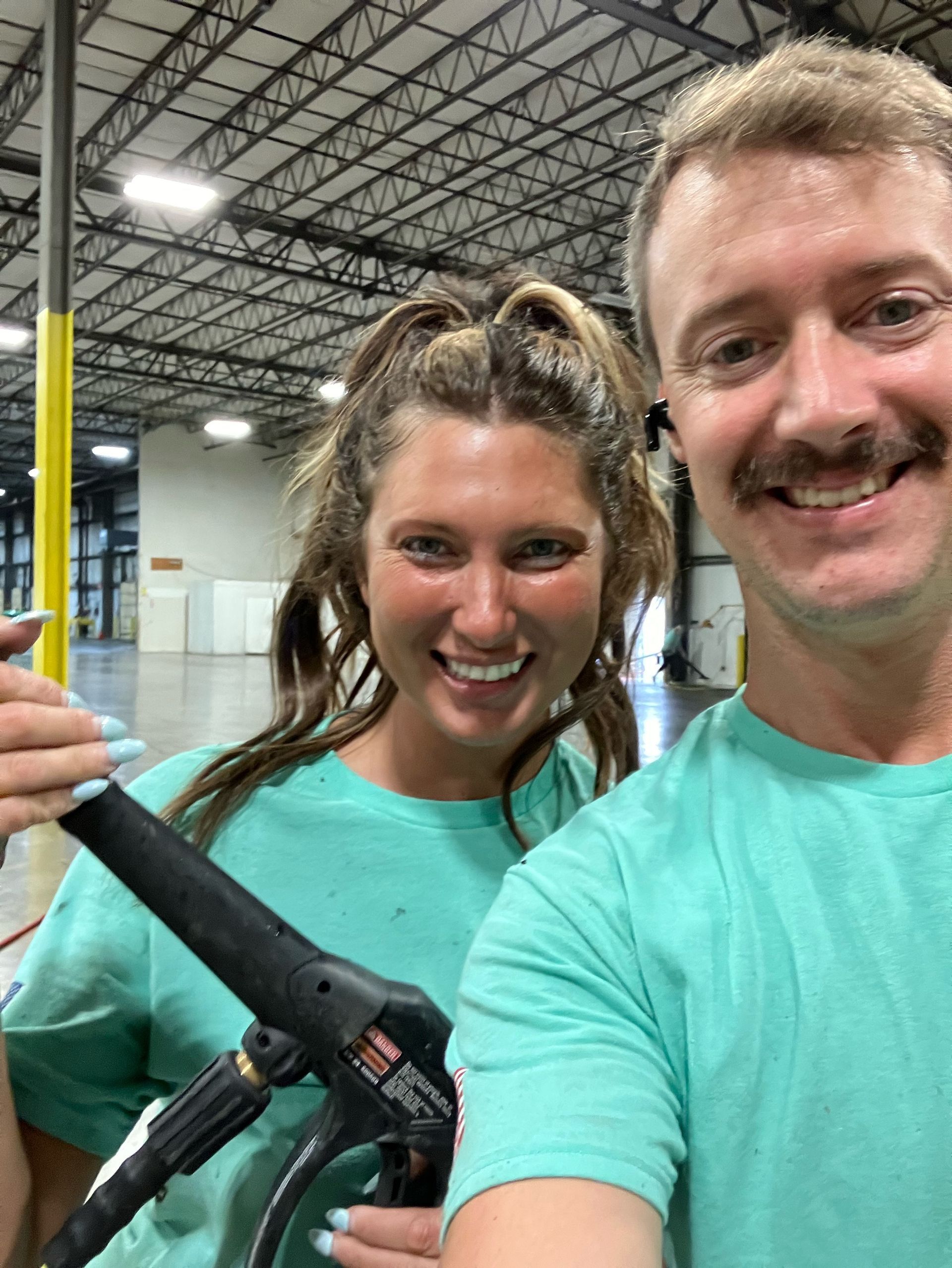 Two people smiling in teal shirts, one holding a pressure washer, in a warehouse setting.