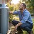 Man inspecting an air conditioning unit outside, squatting with tools, in front of a house, sunny day.