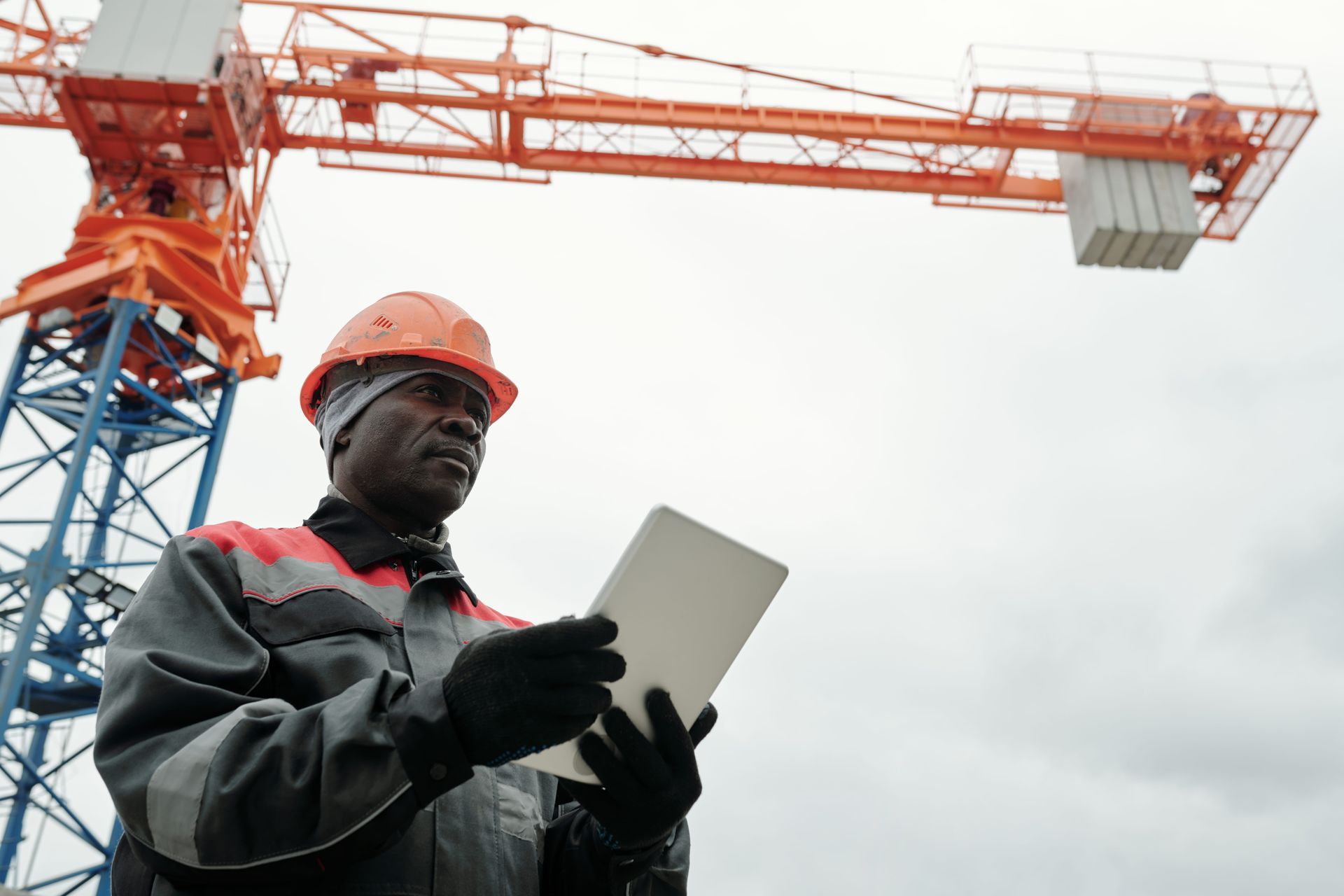 Construction worker in orange hard hat using a tablet beside a tower crane