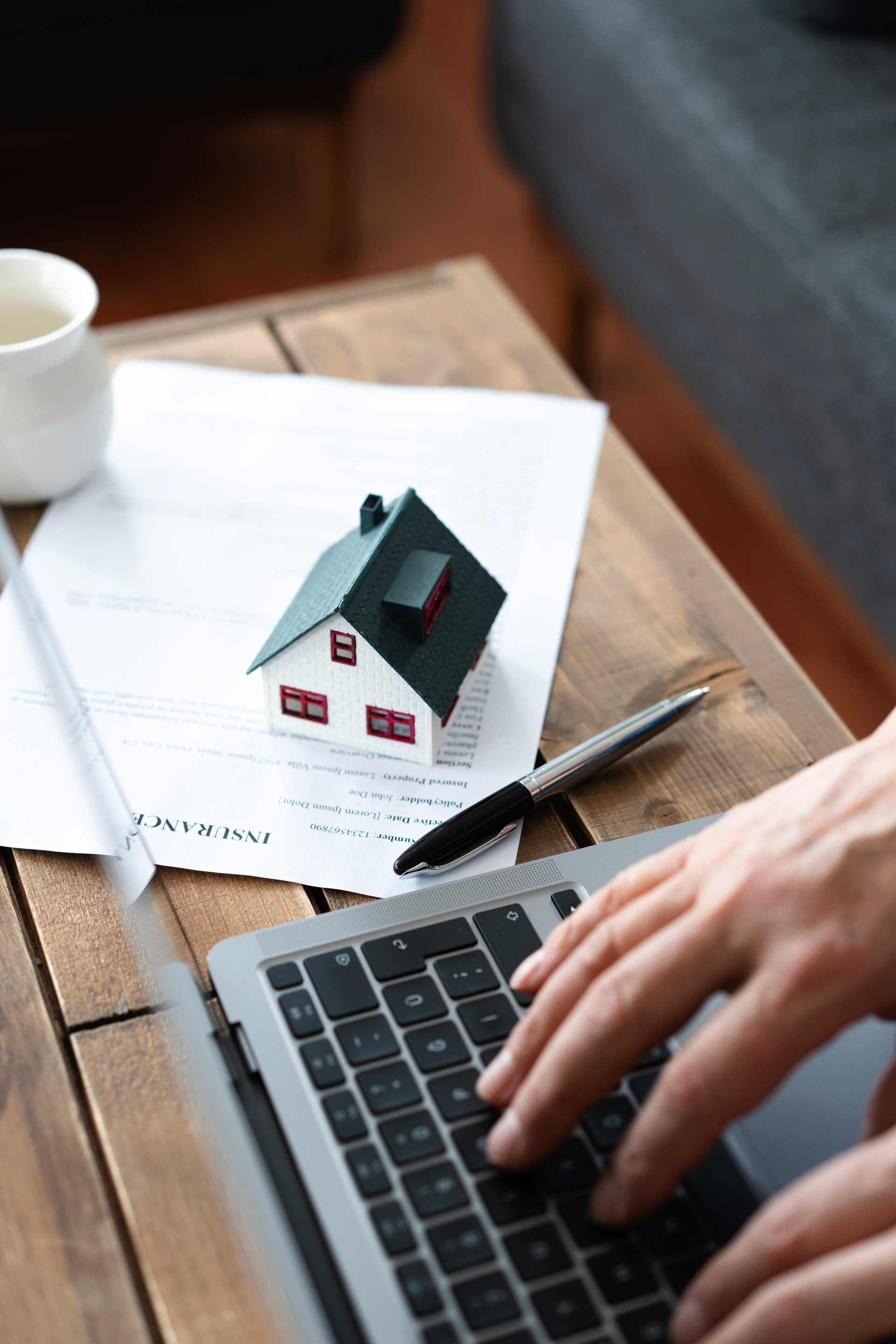 Person typing on laptop, house model and documents on a wooden table, suggesting real estate or home buying.