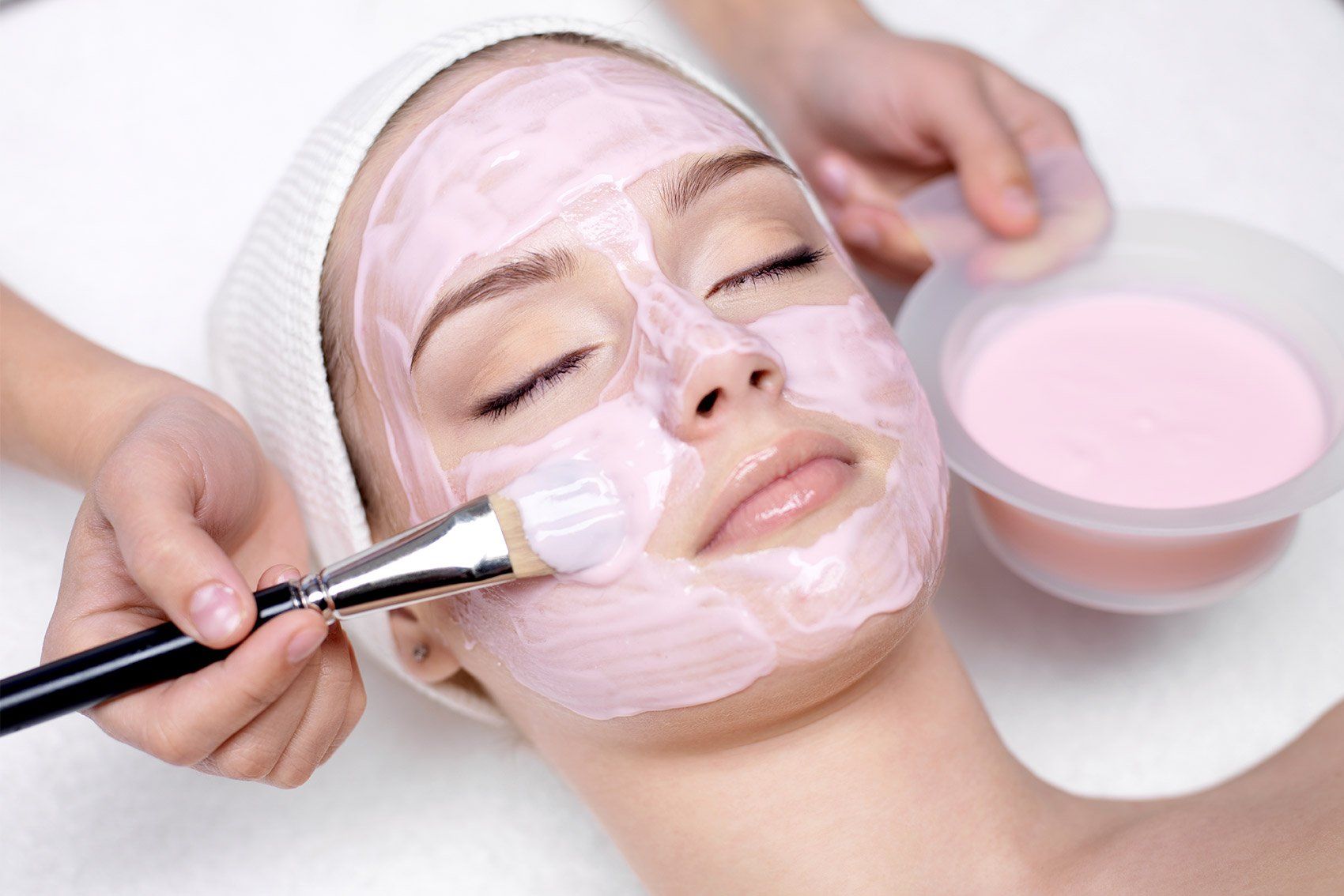 Cosmetic facial mask being brushed onto a woman’s face, with a bowl of pink cream nearby