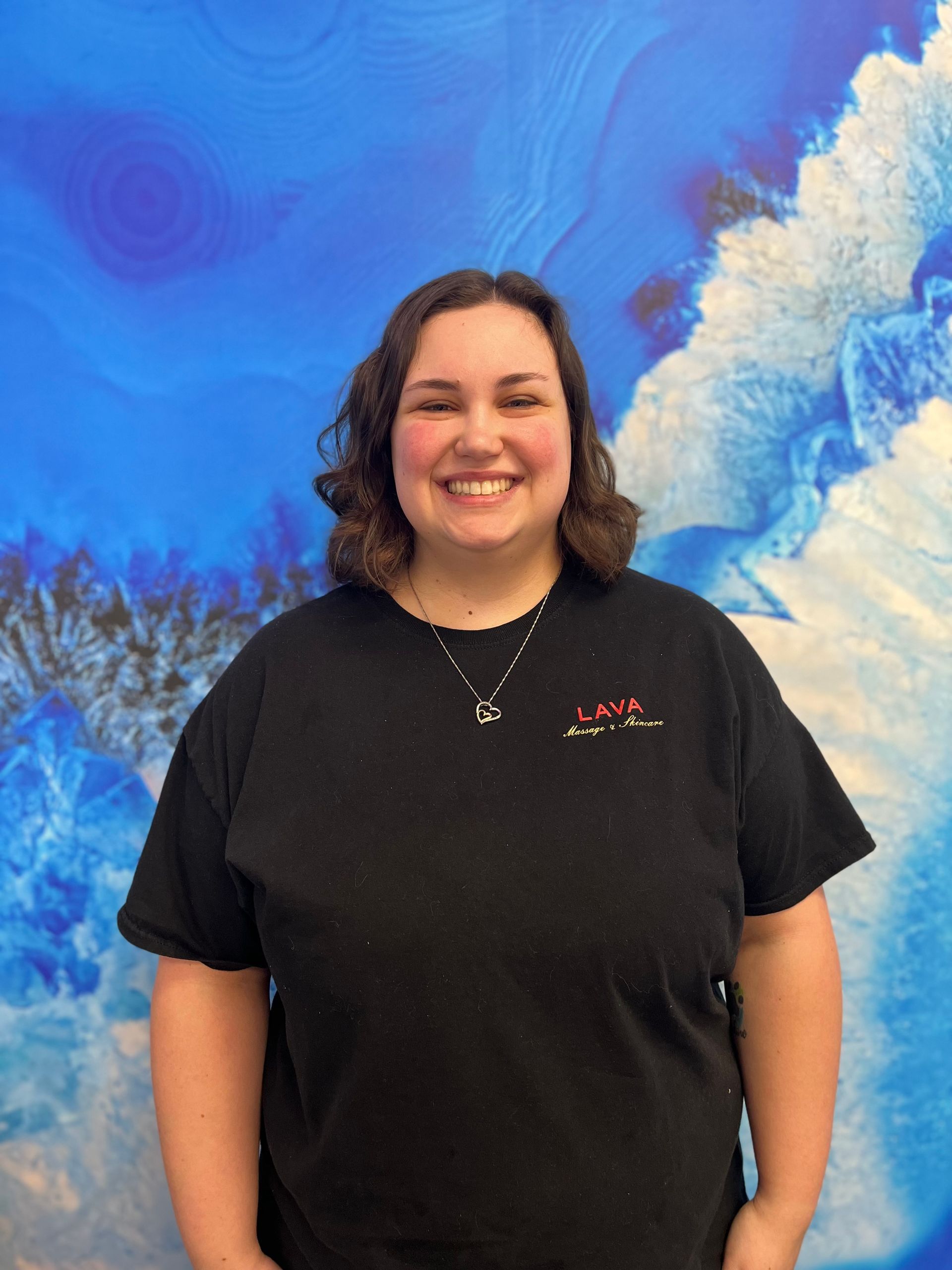 Smiling person in a black shirt standing before a bright blue cloud-like mural