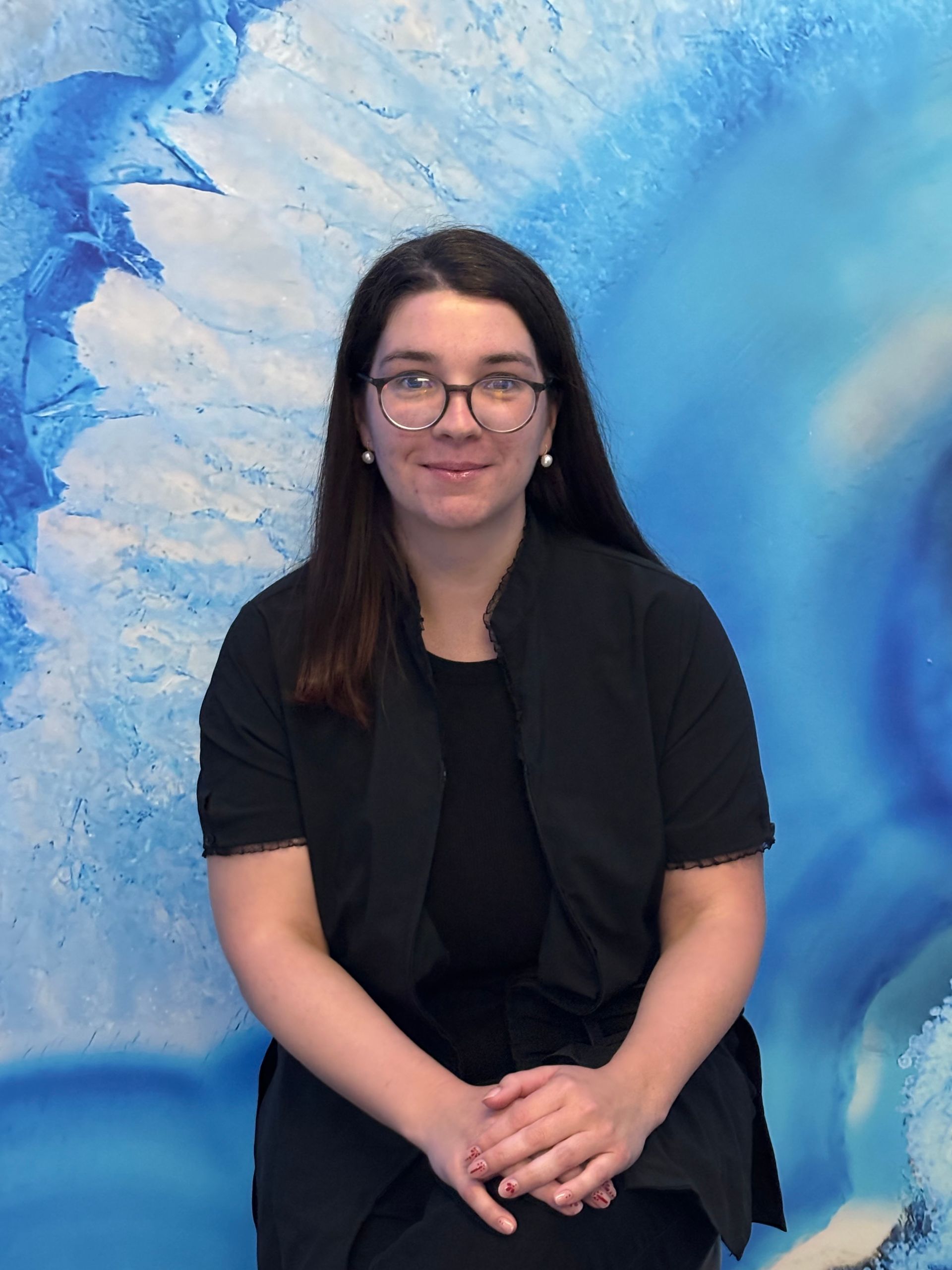 Person seated in front of a blue abstract mural, wearing black and smiling gently