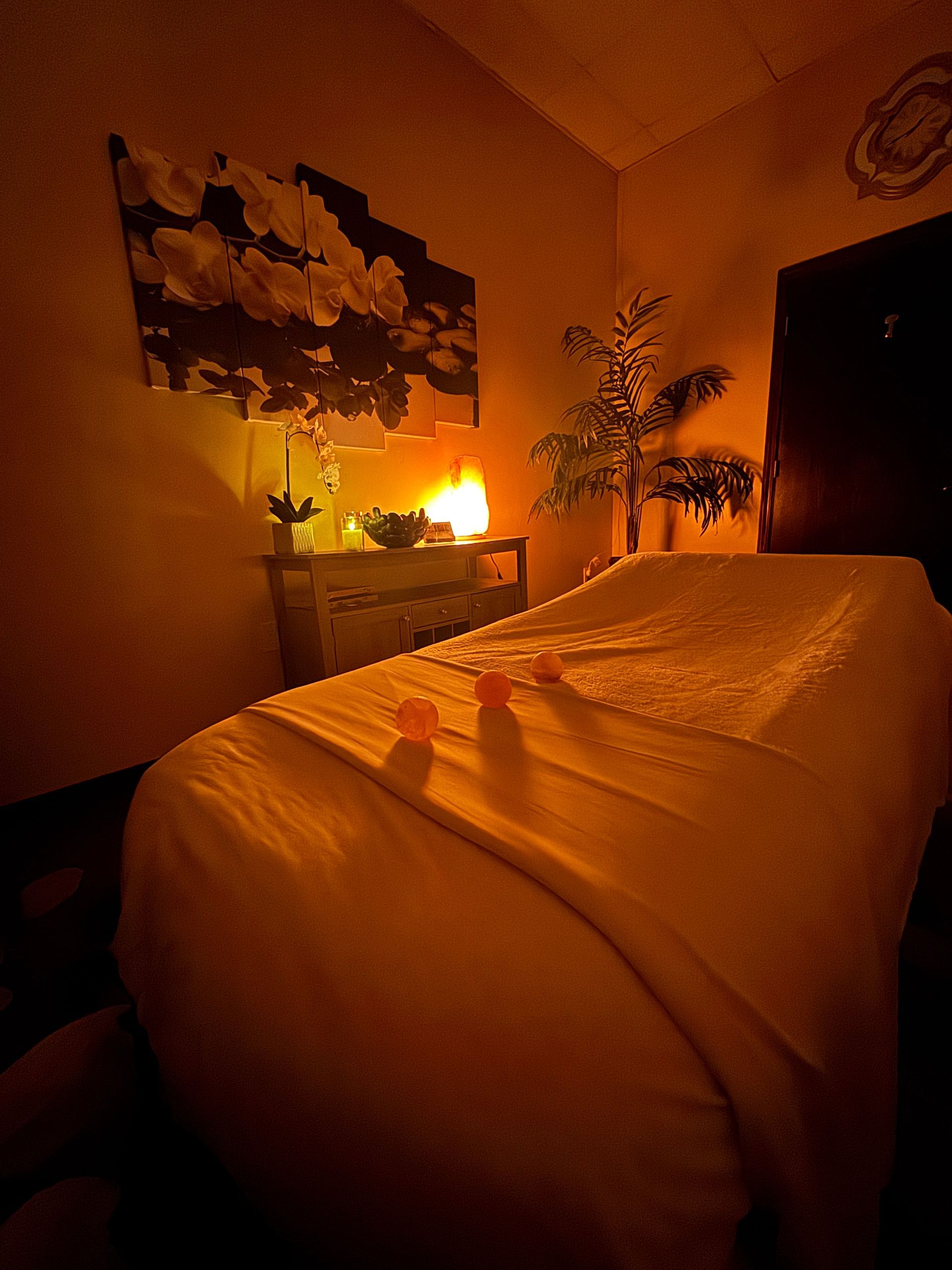 Dimly lit bedroom with a made bed, warm lamp glow, wall art, and a plant.