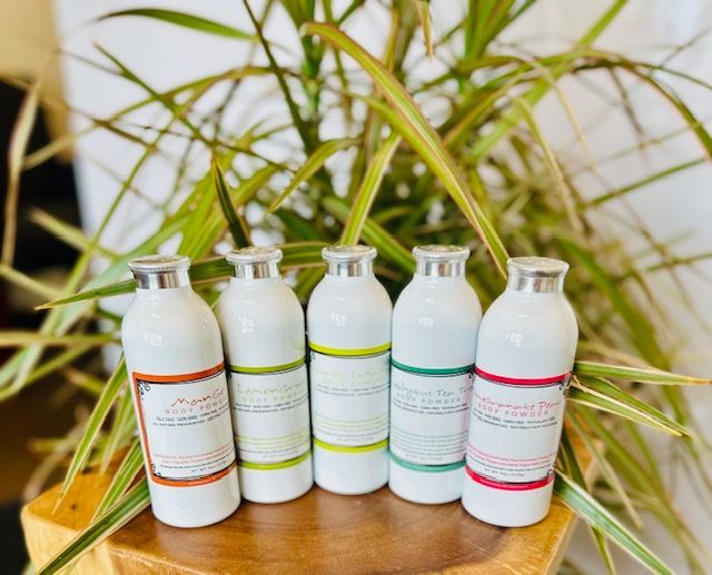 Five small white bottles with colored labels arranged on a wooden tray against leafy green background.