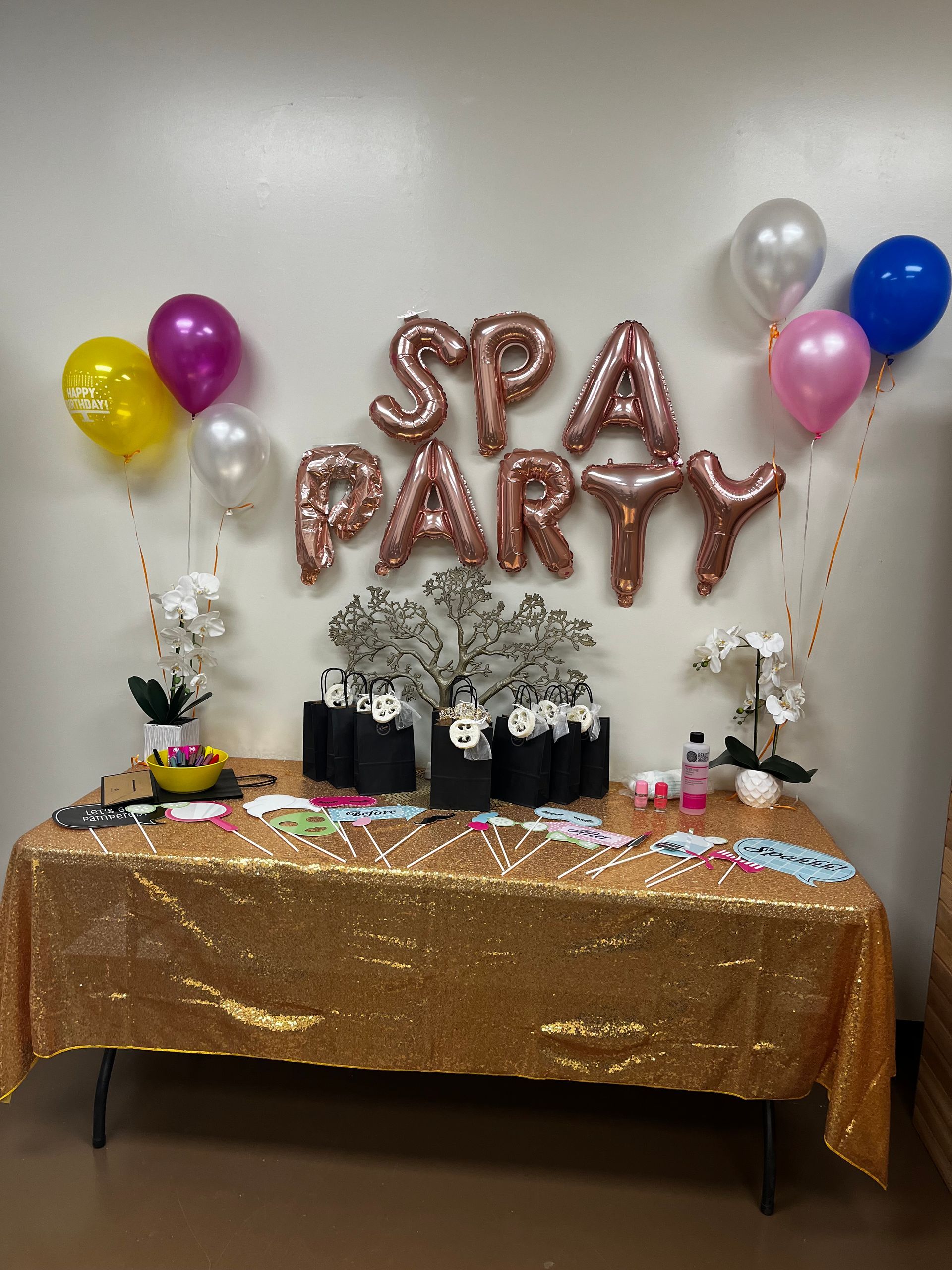 Spa party table with gold sequin tablecloth, balloons, gift bags, and pink balloon letters on a wall