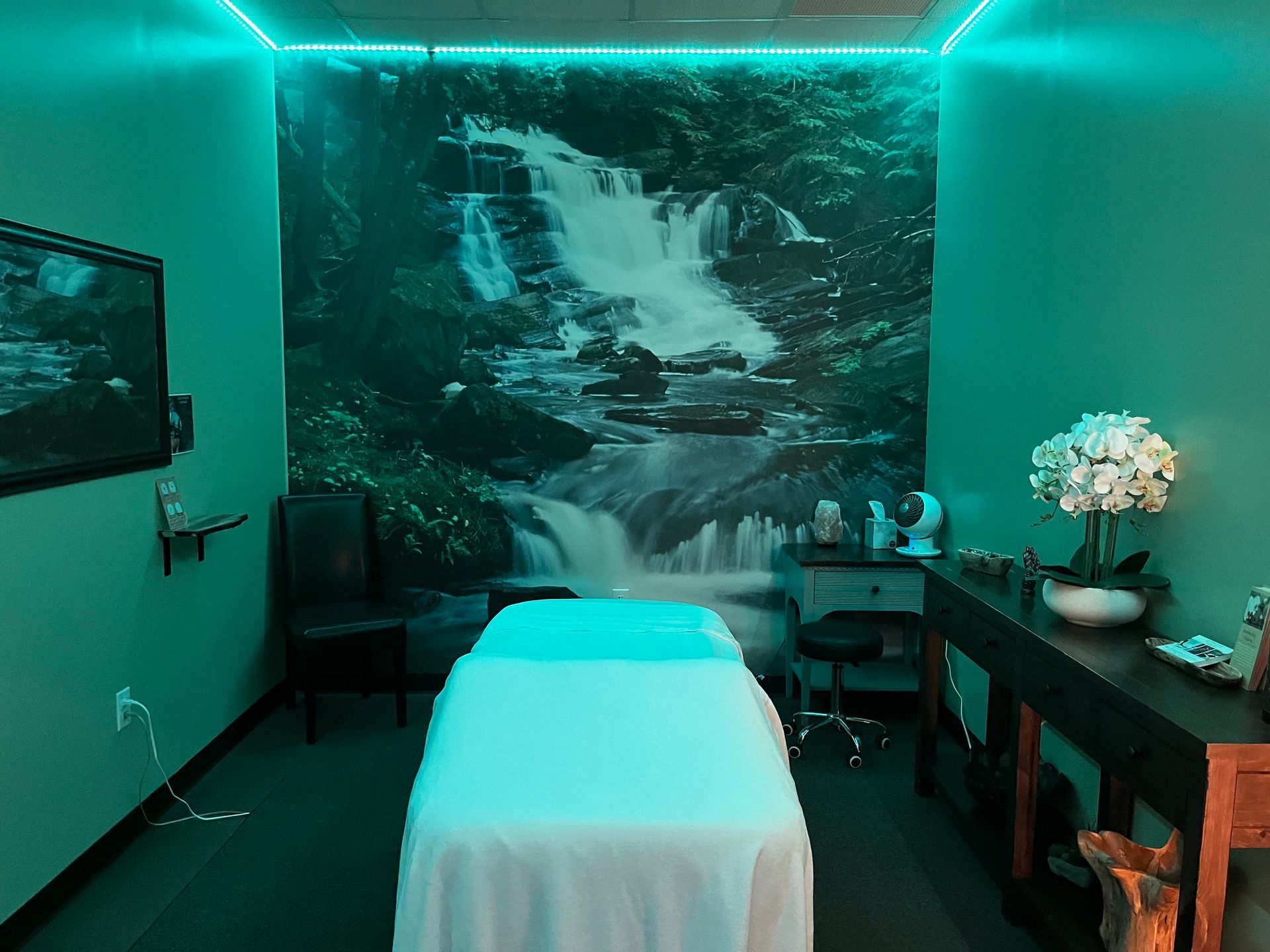 Teal-lit massage room with a treatment table, wall waterfall mural, and side table with flowers.