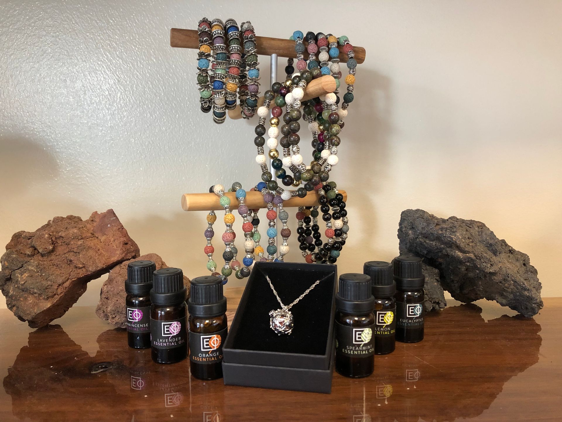 Jewelry display with beaded bracelets, necklaces, rings, and small bottles on a wooden surface, flanked by rocks