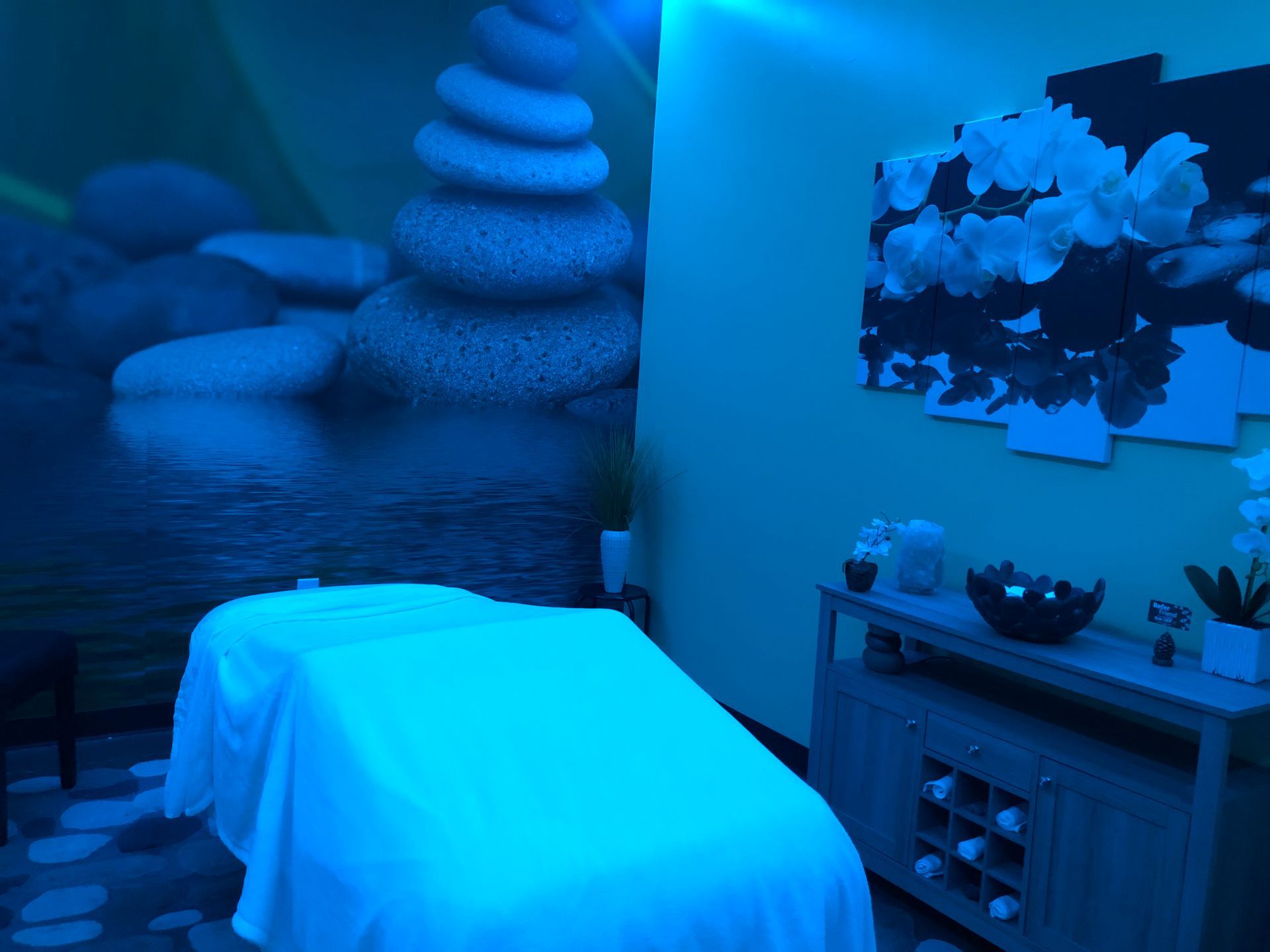 Blue-lit massage room with a treatment table, stacked stones mural, and shelf with spa items