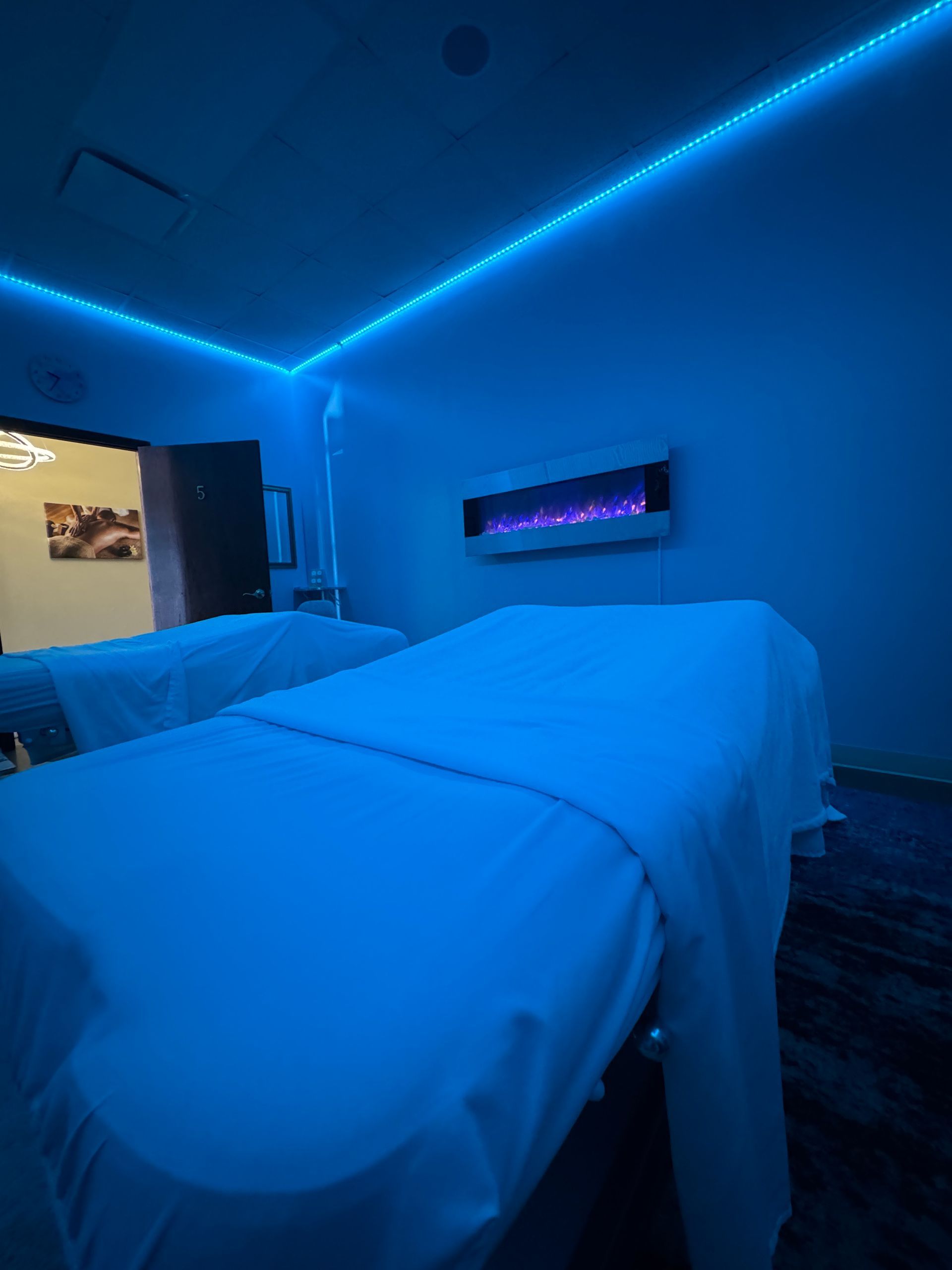 Blue-lit bedroom with an unmade bed, wall-mounted TV, and open doorway to a lit hallway.