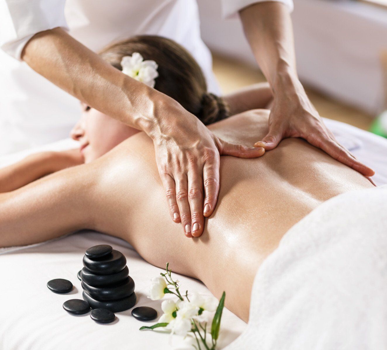 Spa massage with hands pressing a woman’s oiled back, black stones and white flowers nearby