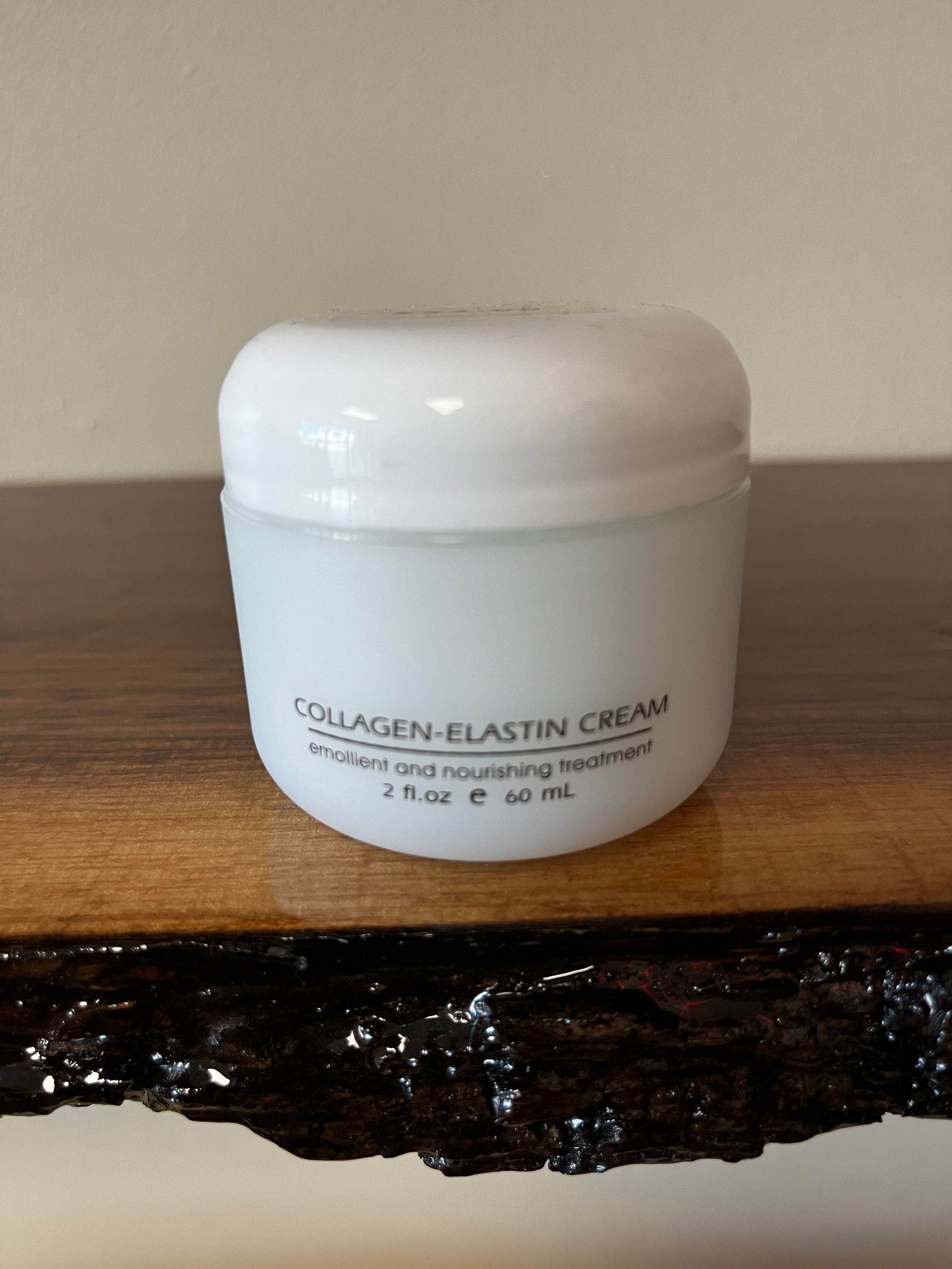 White cosmetic jar labeled “Collagen-Elastin Cream” on a wooden surface against a beige wall.