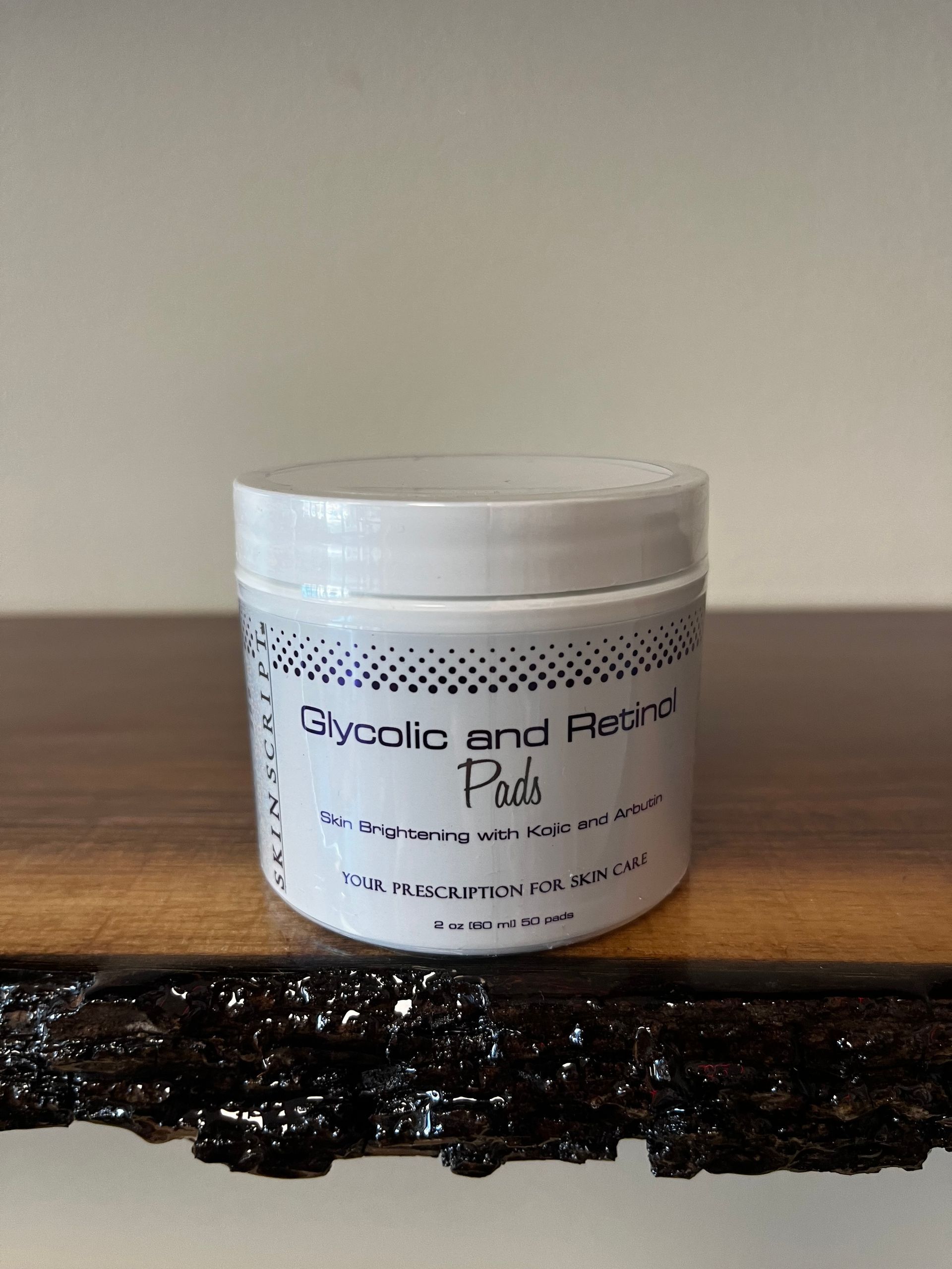 White jar labeled “Glycolic and Res…” on a dark wooden surface against a plain wall.