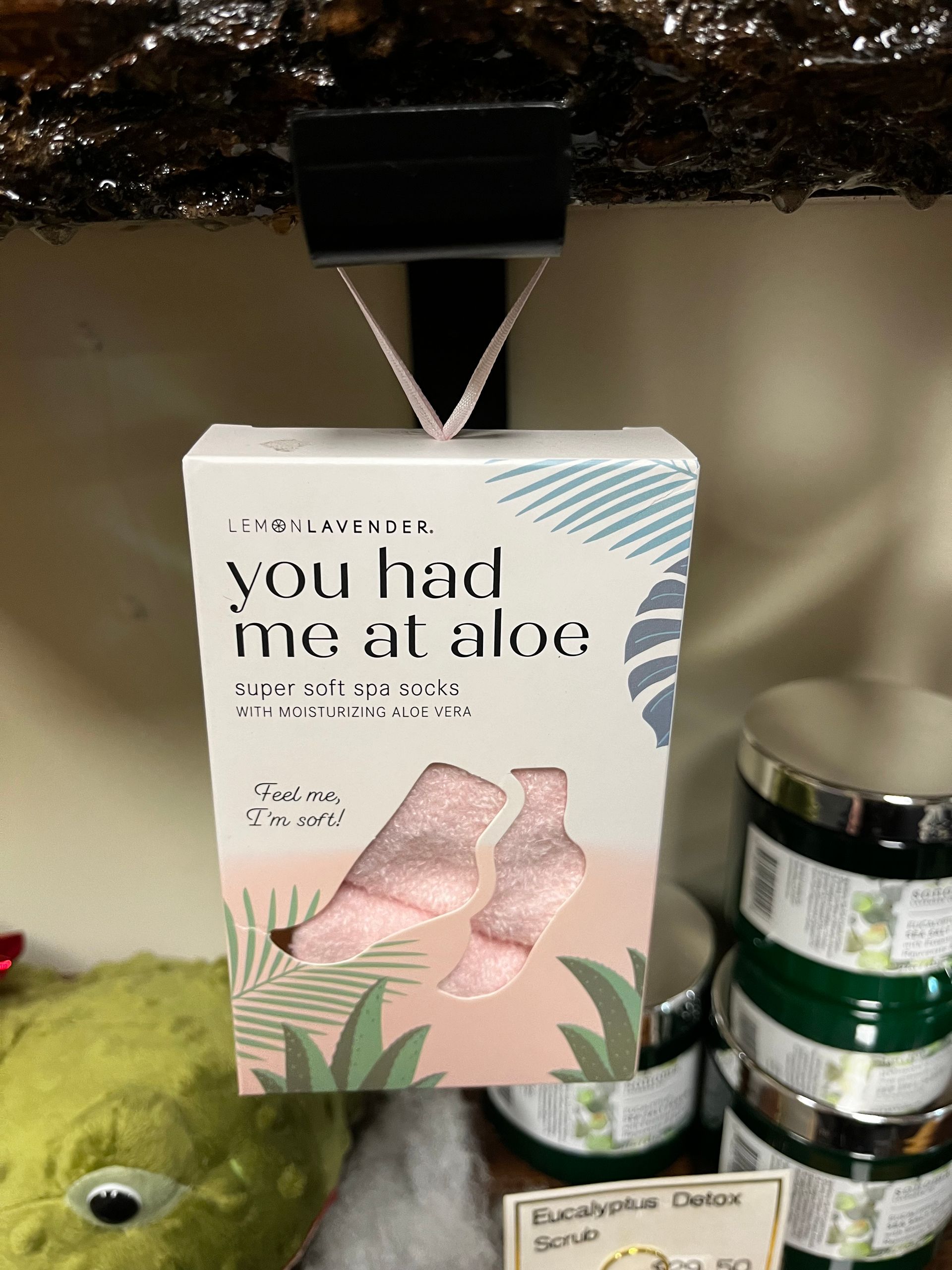 Box of “you had me at aloe” product displayed on a shelf, with green plush toy and jars nearby.