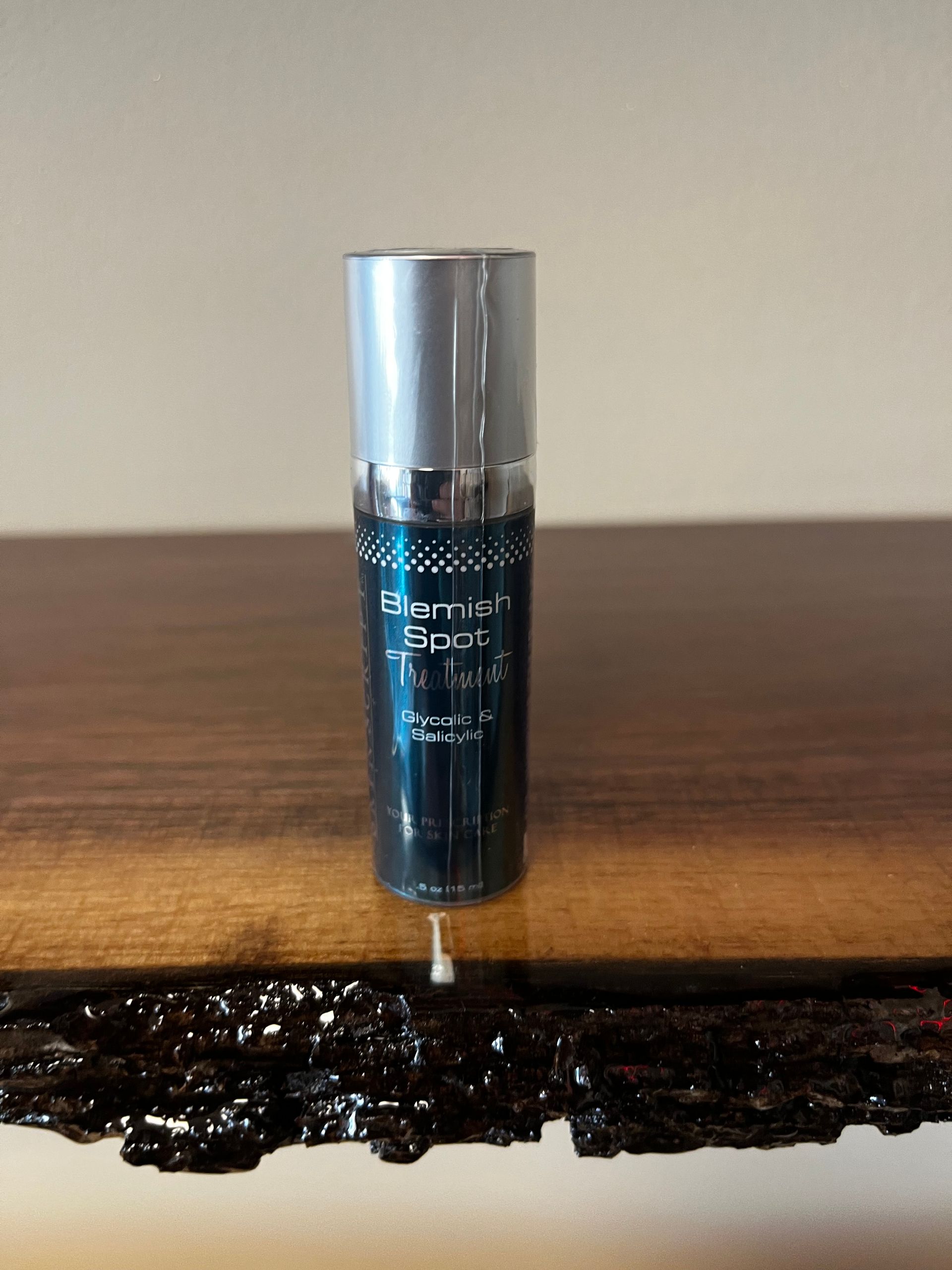 A dark blue skincare bottle with a silver cap standing on a wooden table against a beige wall.