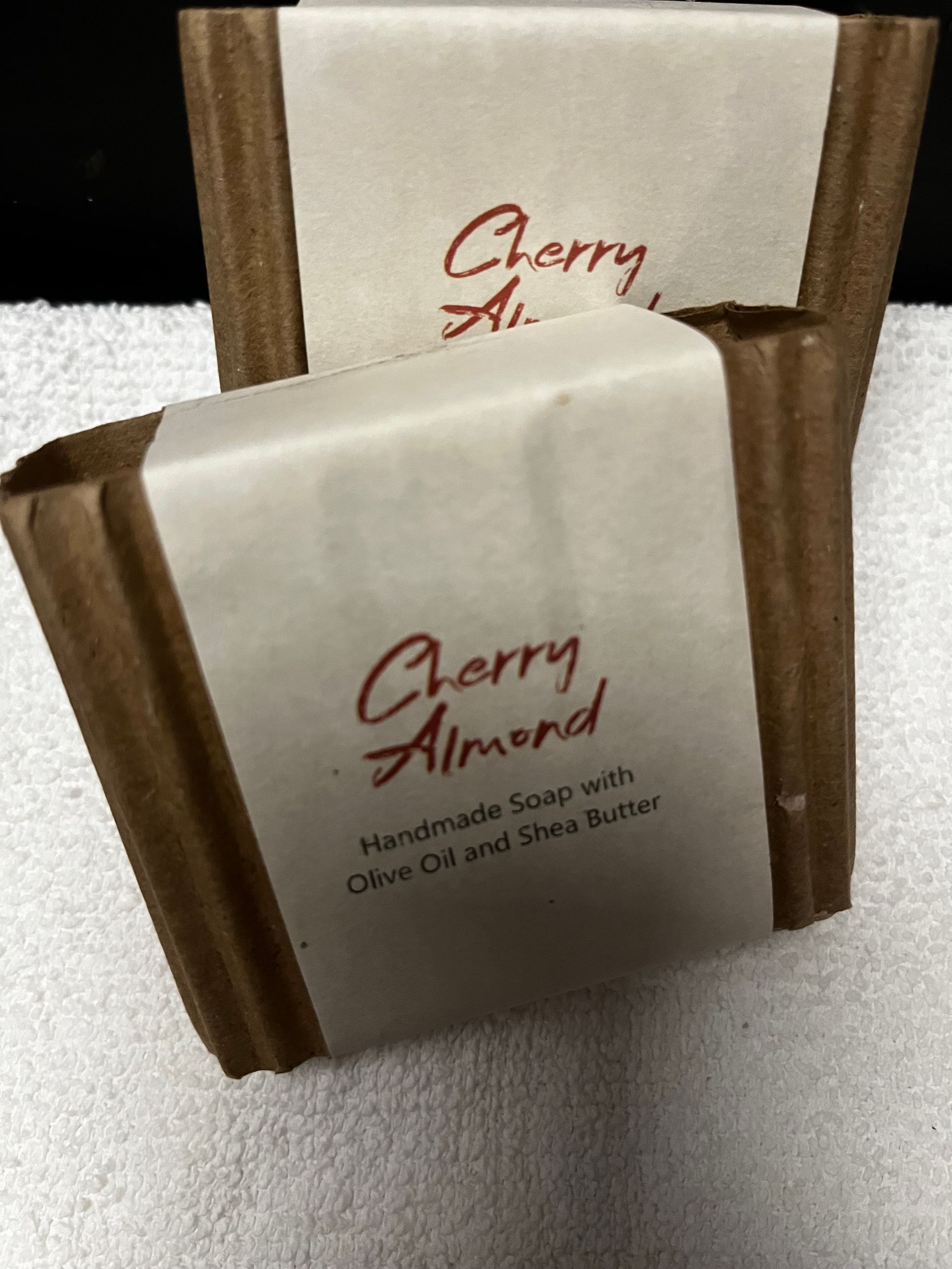 Two Cherry Almond soap bars in white wrappers with brown sides, stacked on a soft gray surface.