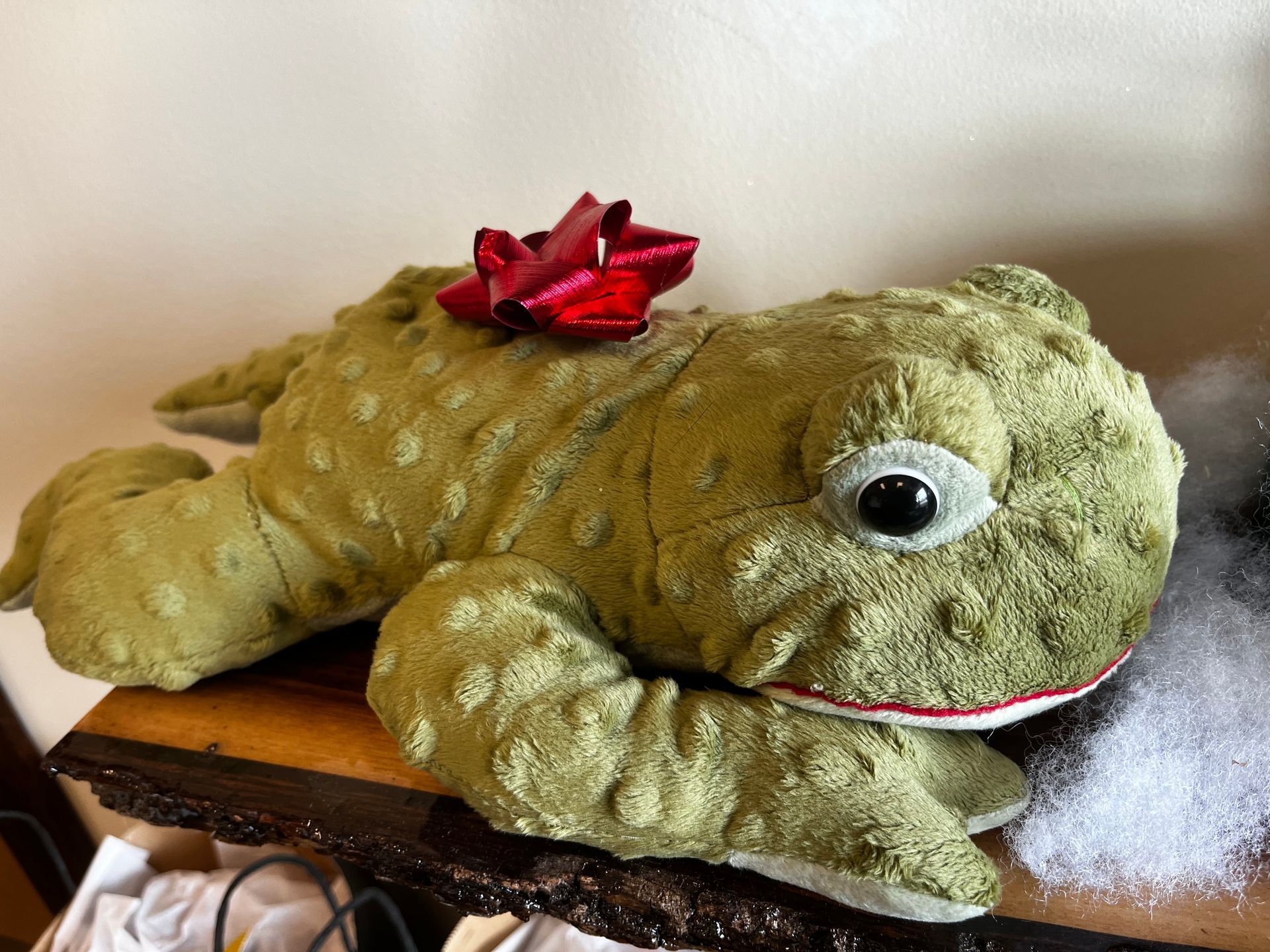Green plush frog with a red bow on a wooden surface, next to white fluff