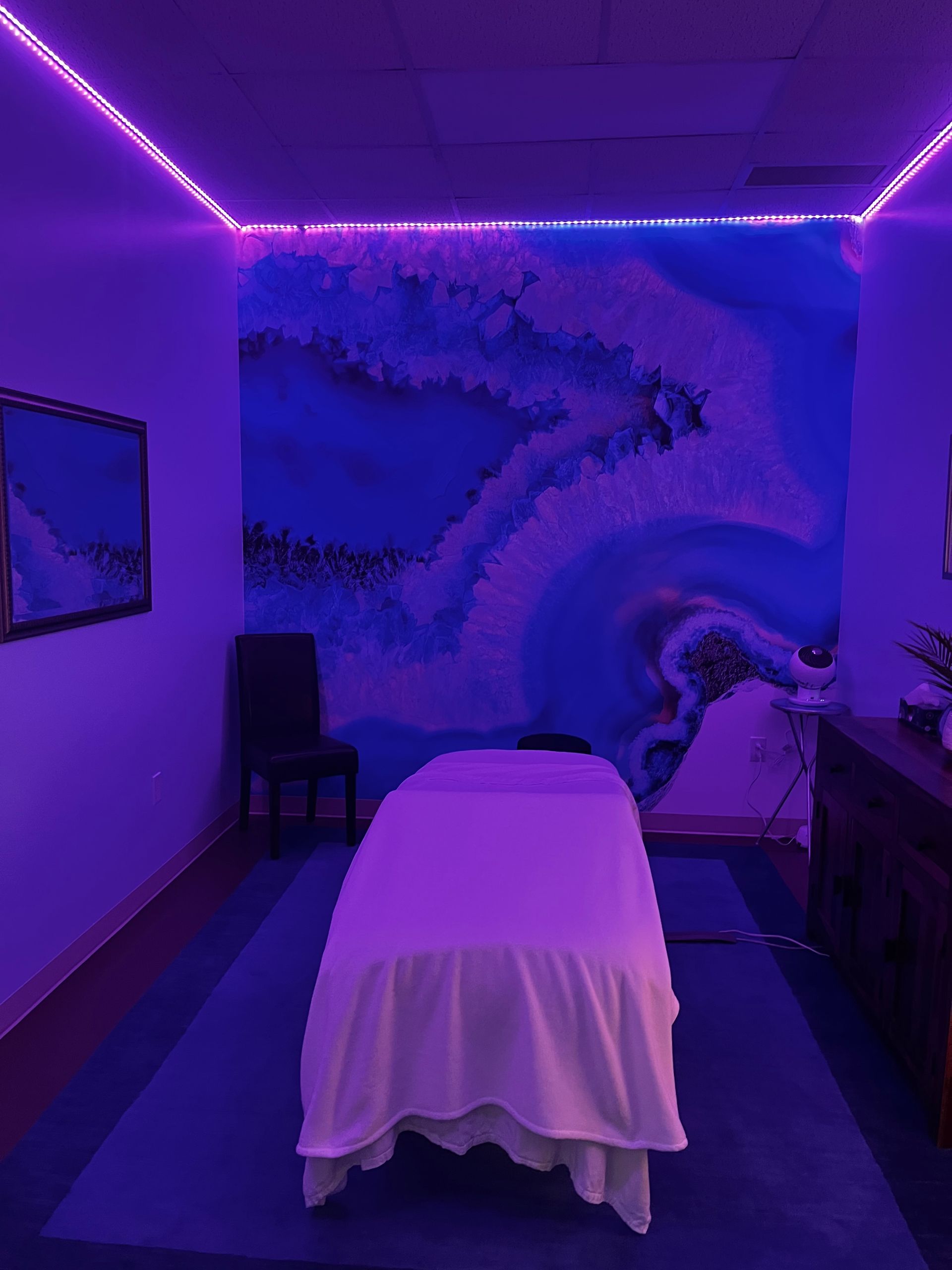 Purple-lit massage room with a treatment table draped in white, mural wall, and LED ceiling lights