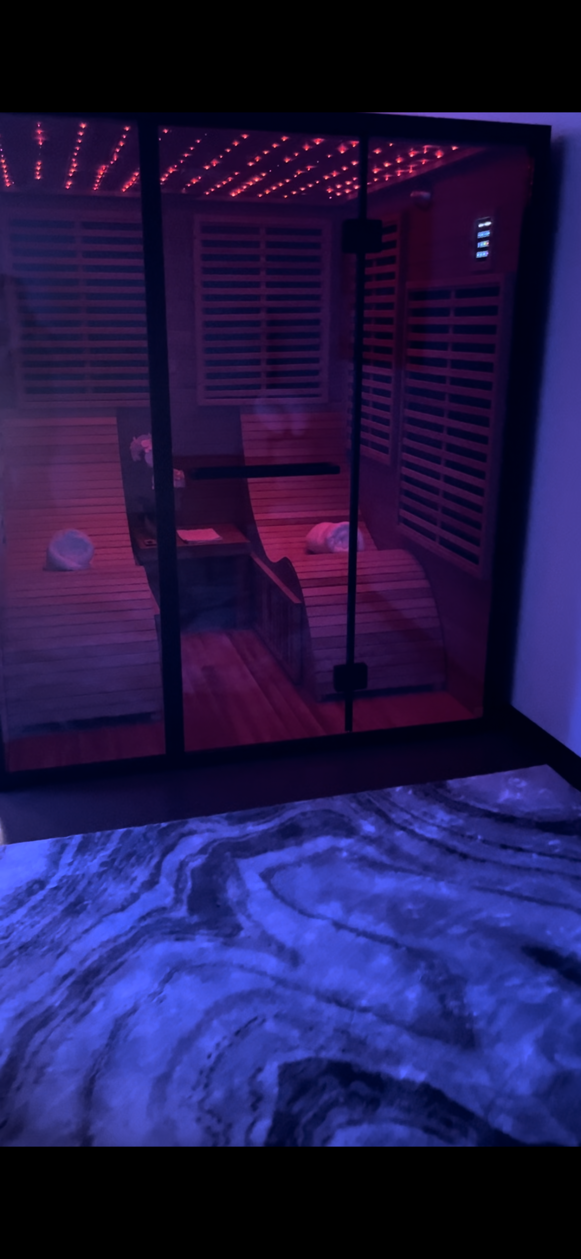 Purple-lit glass shower stall with bench and tiled walls, seen from a bedroom with a blue rug.