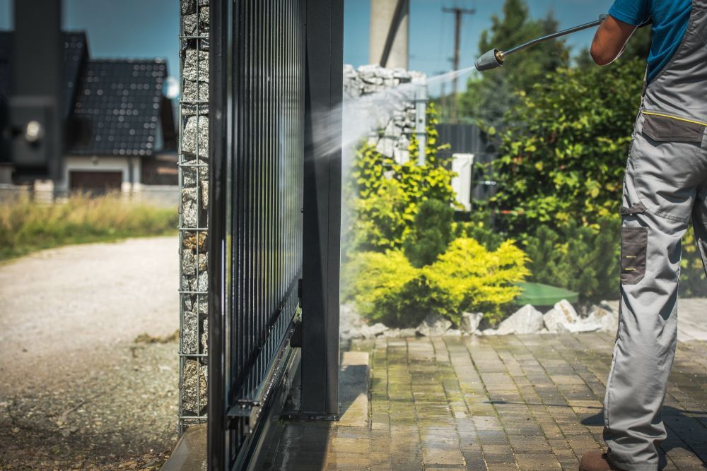 Person Power Washing A Dark Metal Gate On A Brick Patio; Plants And A House Are In The Background — Rocky Commercial Cleaners In Berserker, QLD