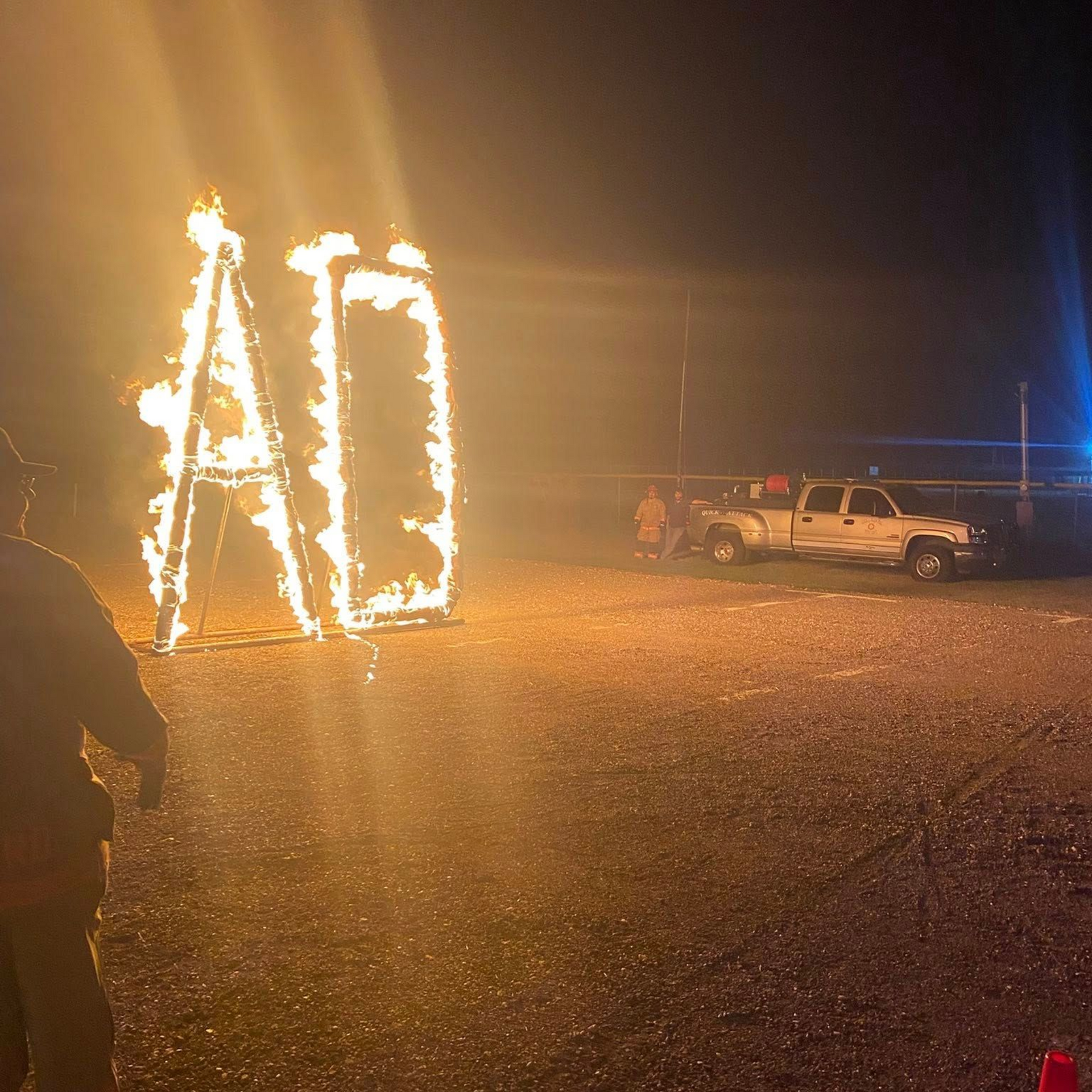 Flaming “AO” letters lit up at night near parked vehicles on a gravel lot.