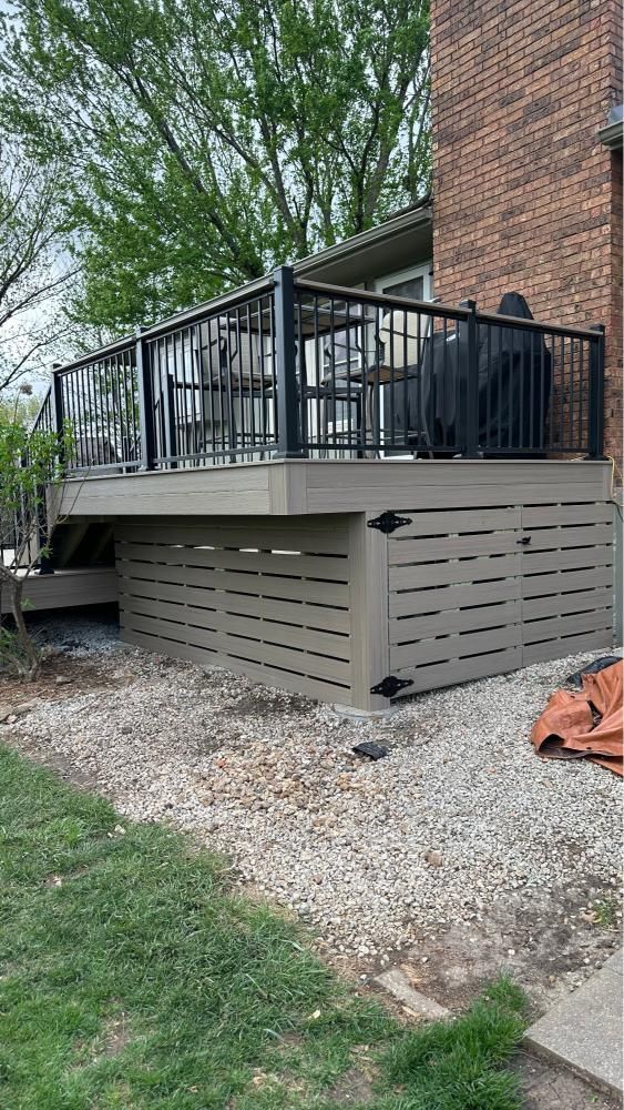 Back deck with black railing on a brick house above a gray louvered enclosure and gravel ground