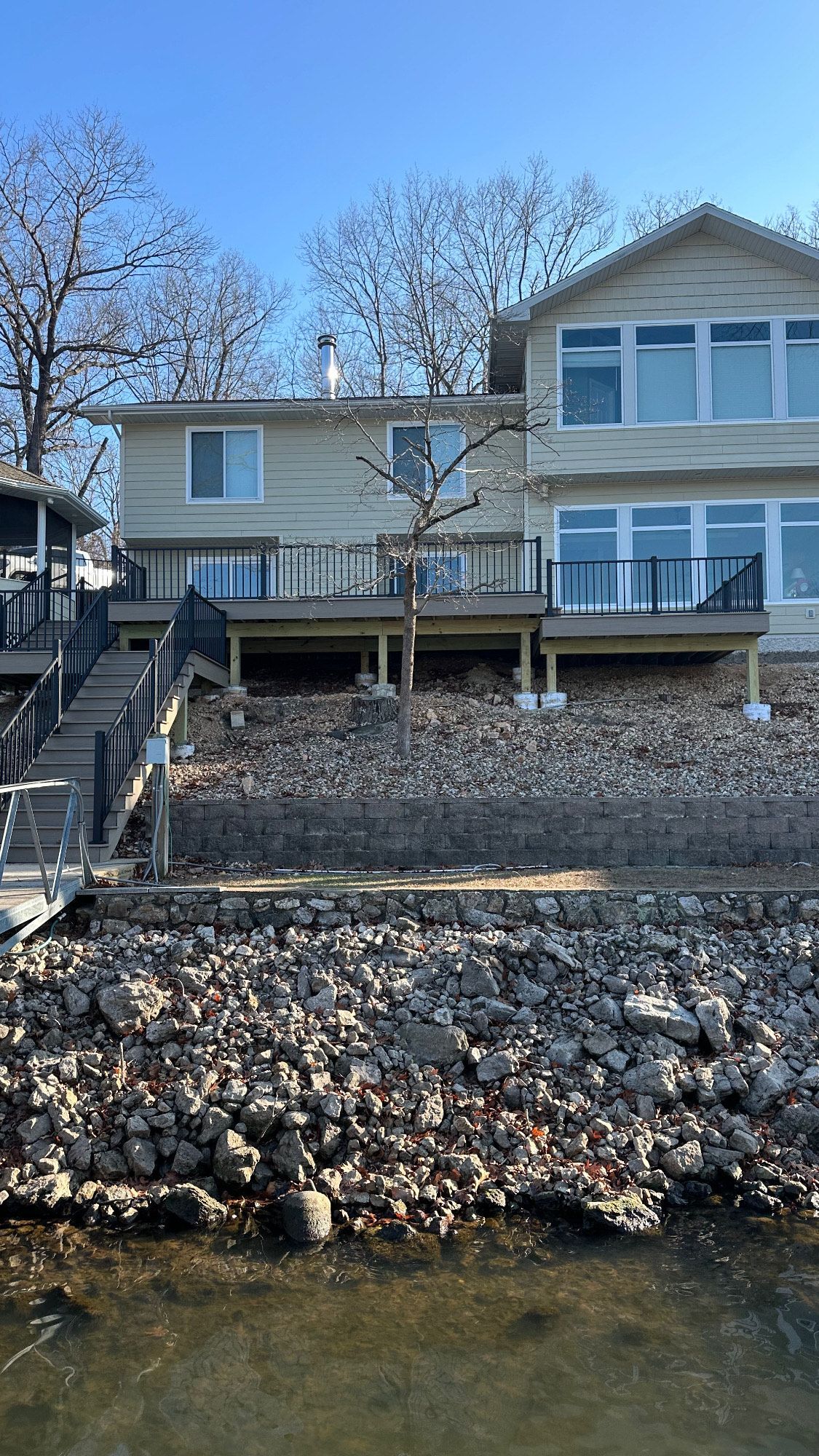 Two-story lakeside house with a deck, rocky shoreline, and bare trees on a sunny day