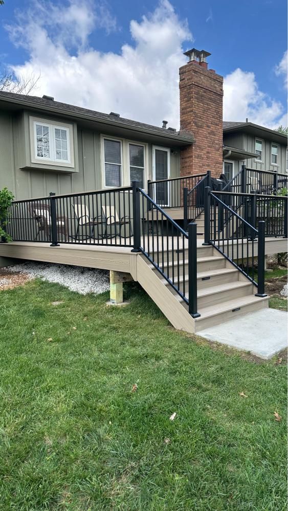 Front porch with black railings and stairs, attached to a gray house with a brick chimney and lawn.