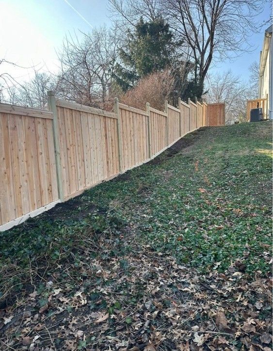 Wooden privacy fence along a grassy, leaf-covered slope with trees and blue sky in the background