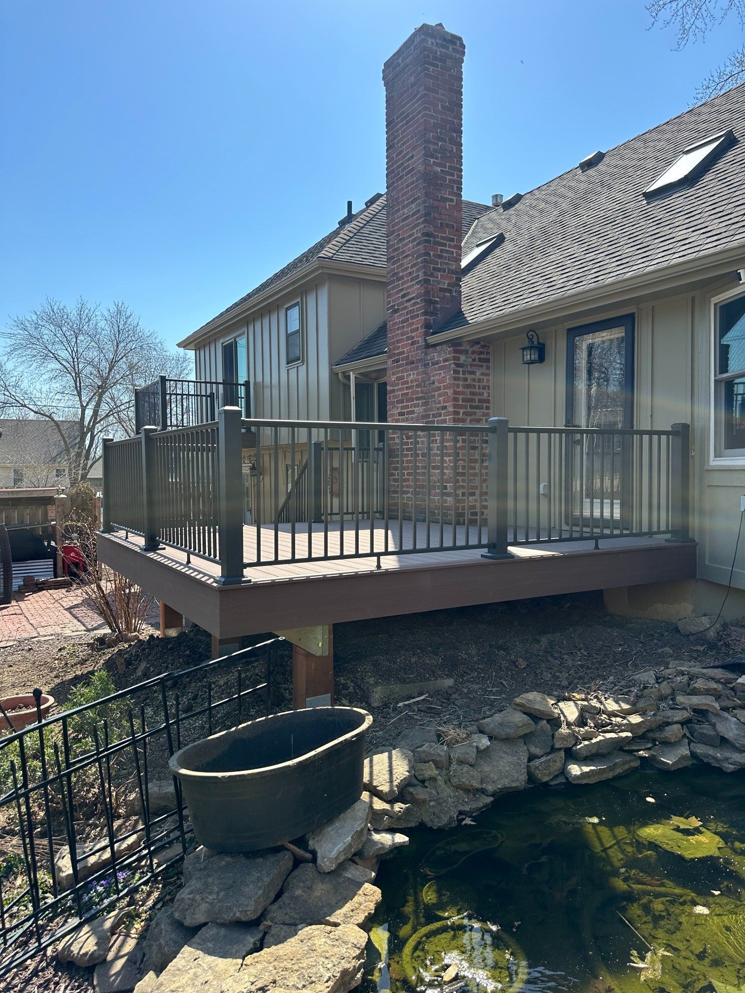 Back deck with metal railing, brick chimney, and a pond beneath a house on a sunny day