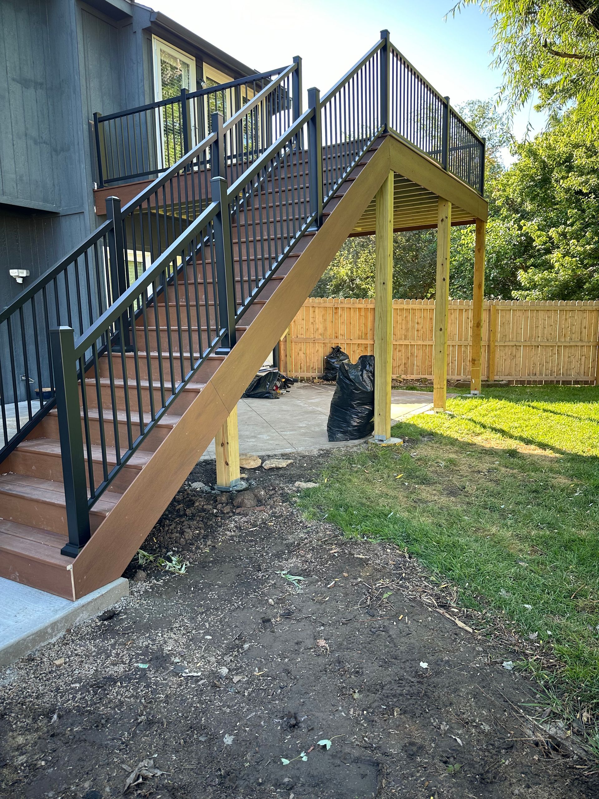 Outdoor wooden staircase with black railings beside a house, leading to a raised deck over a grassy yard.