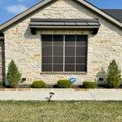 Tan stone house with a large window covered by a black awning, small bushes and trees in front.