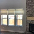 Three windows with shades, wooden fence outside, brick fireplace to the right.