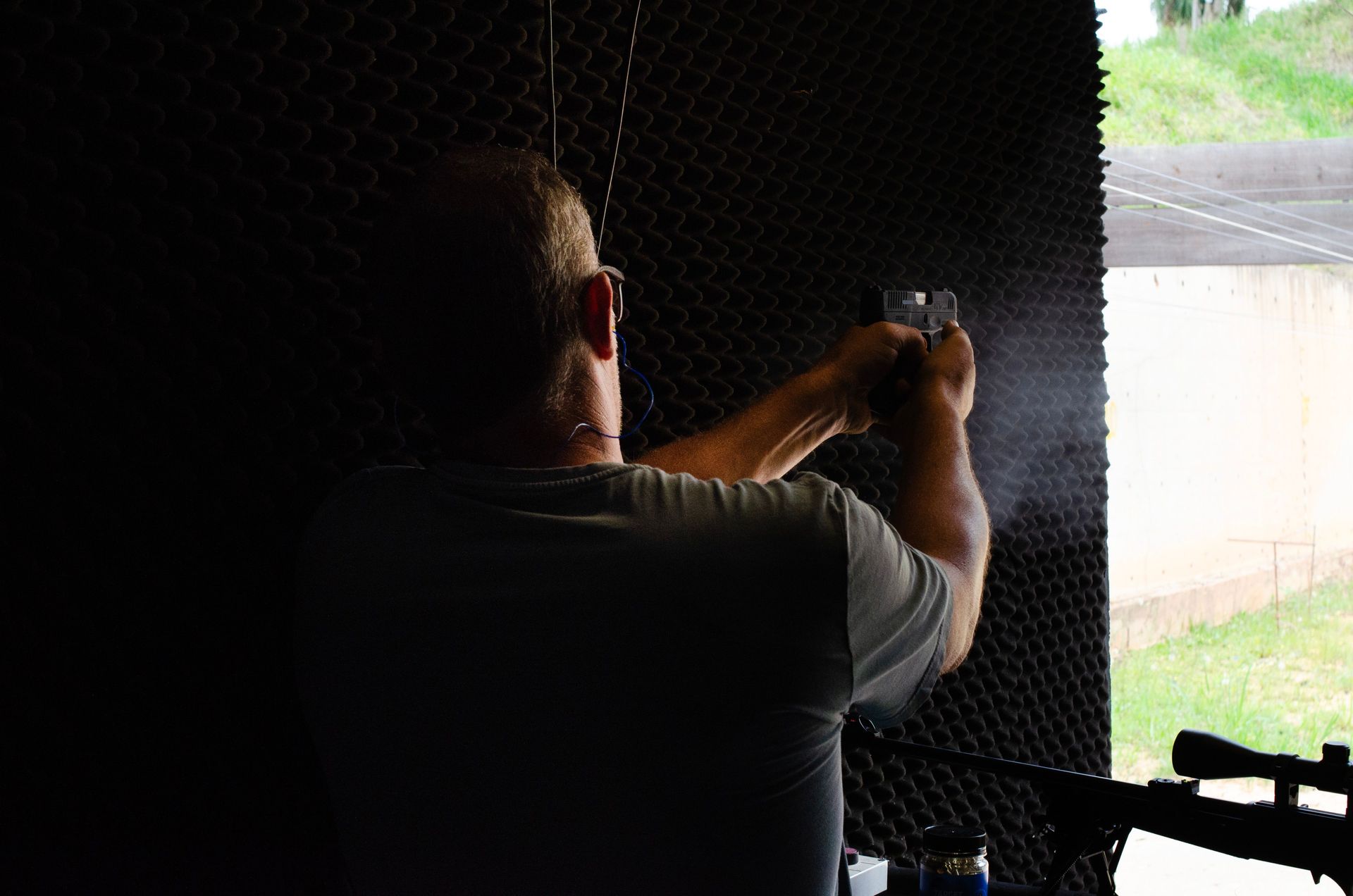Person aiming a handgun at an outdoor range target through a shooting booth opening