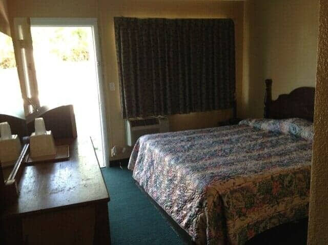 Hotel room with two beds, a desk, and an open door to a bright balcony or patio.