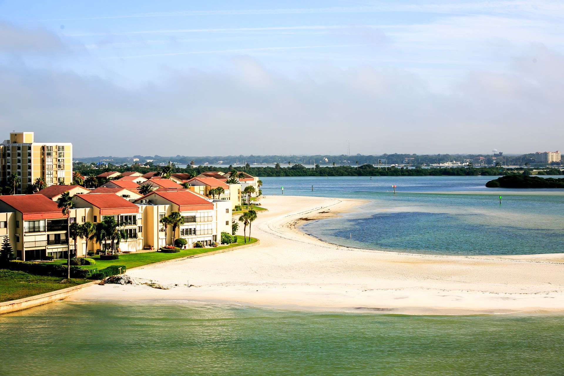 Coastal resort with red-roofed buildings beside a sandy beach and blue water under a bright sky